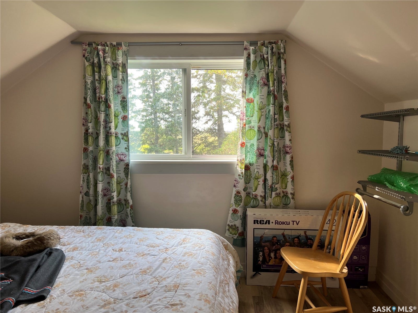 Bright attic bedroom featuring a large double-hung window with natural light, wood-finish flooring, sloped ceiling characteristic of upper-level spaces, and built-in shelving