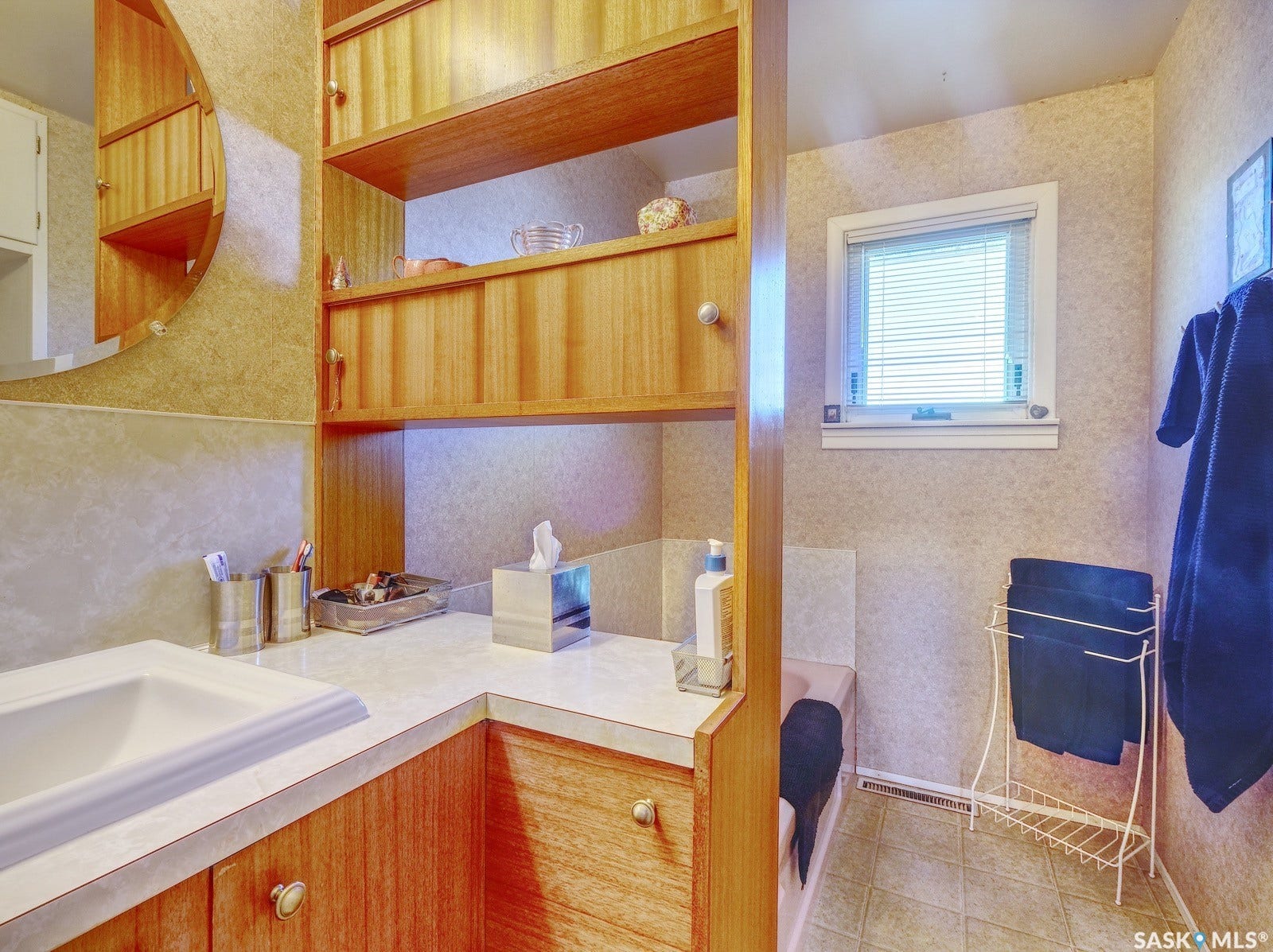 Bathroom featuring a built-in wood-finish vanity with an integrated sink and light-toned countertop, a window with blinds, and wood-finish shelving