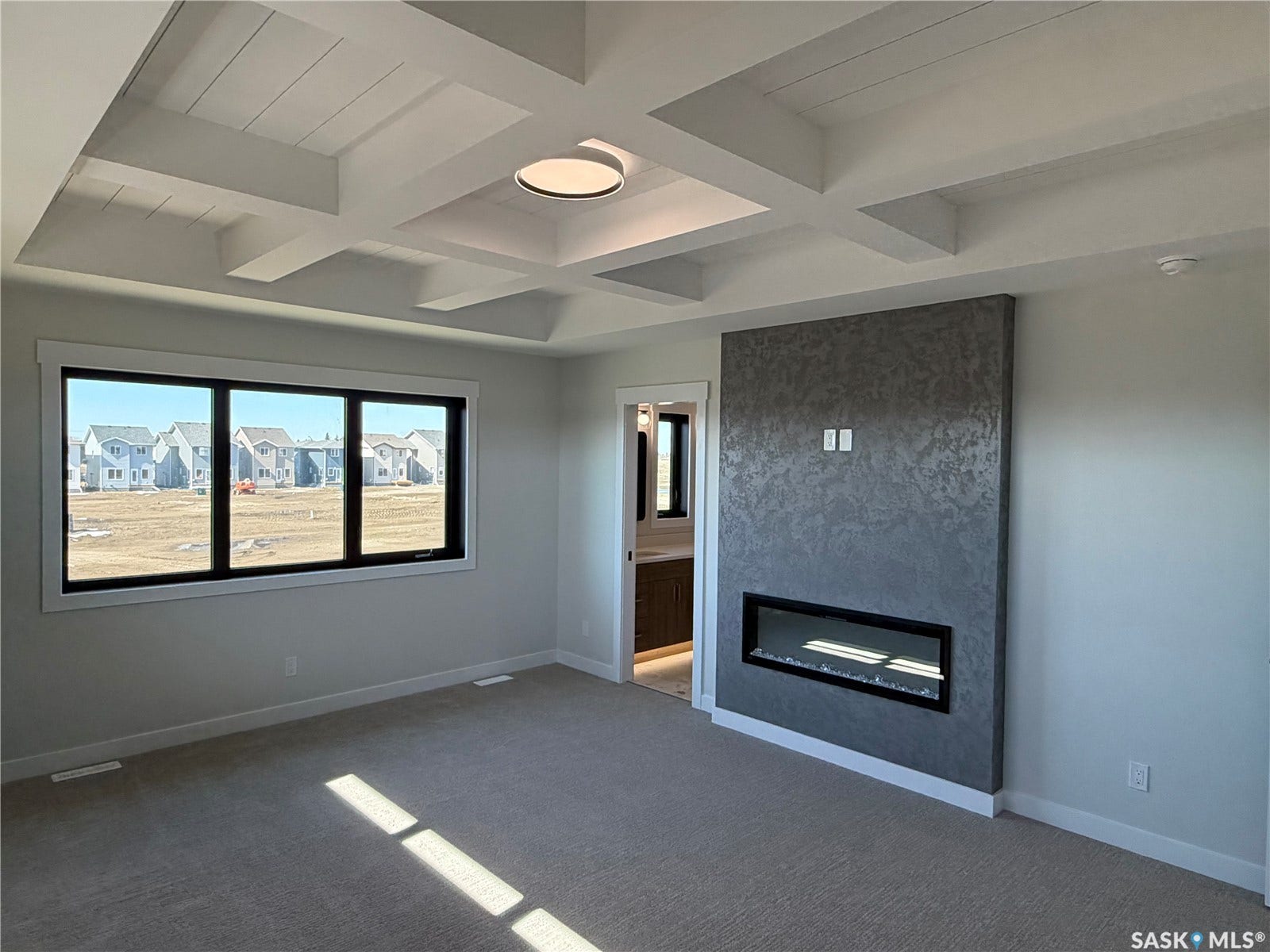 310 Traeger Manor, Saskatoon SK S7V 1Z4 Coffered ceiling with central flush-mount lighting, grey textured accent wall with a built-in linear fireplace, large triple-pane window, and light-toned carpeting