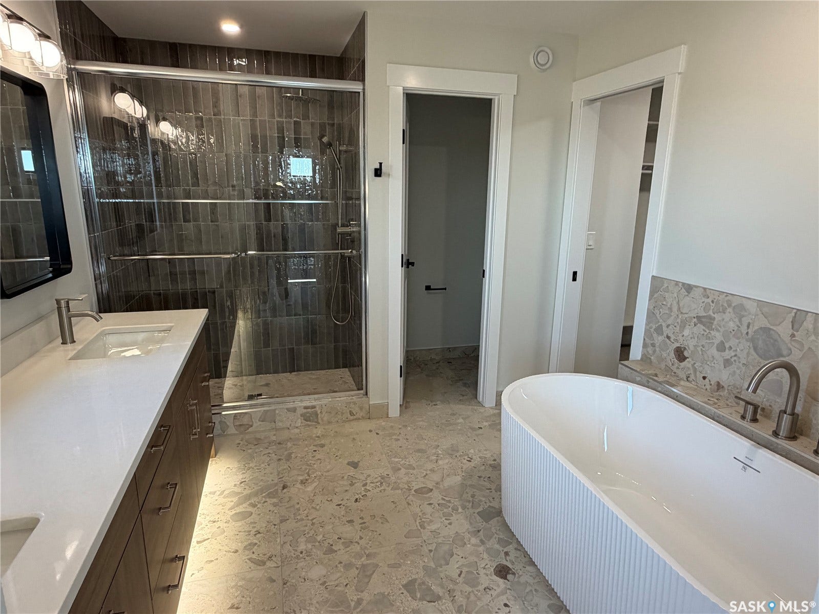 310 Traeger Manor, Saskatoon SK S7V 1Z4 Freestanding tub with fluted exterior, walk-in shower with dark vertical tile, dual vanity with undermount sinks, and light-toned terrazzo flooring