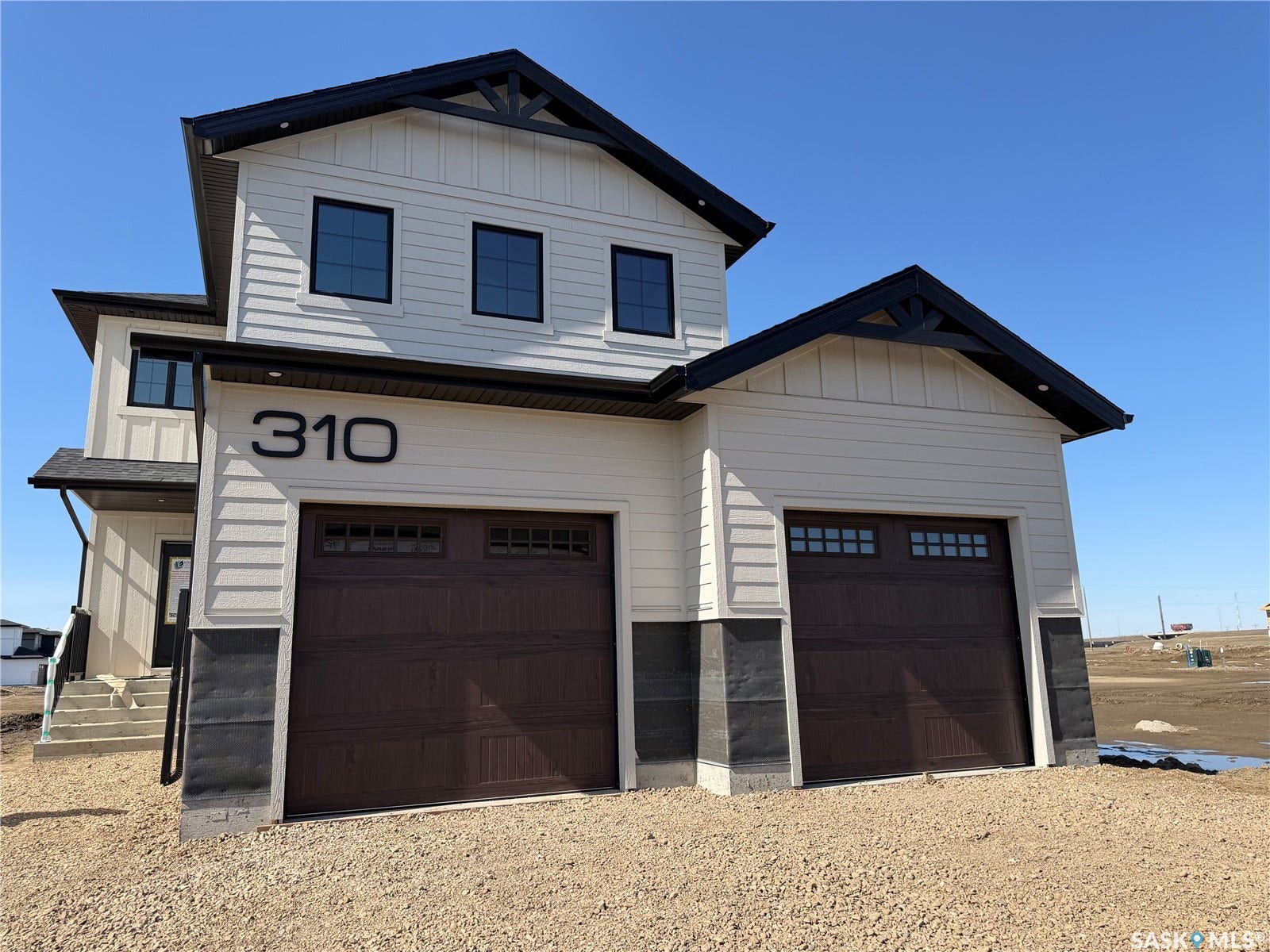 310 Traeger Manor, Saskatoon SK S7V 1Z4 Cream-colored siding exterior with black trim, featuring two dark brown garage doors with upper window panels, and decorative gable brackets