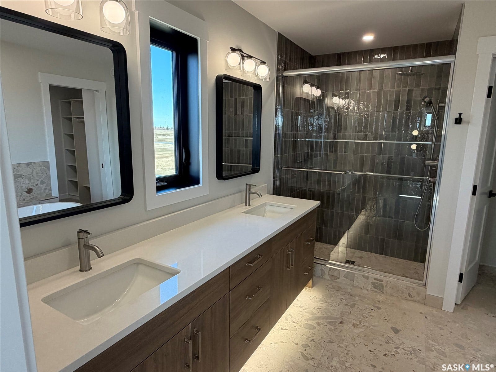 310 Traeger Manor, Saskatoon SK S7V 1Z4 Dual vanities with integrated sinks and white countertops, wood-finish cabinetry, a large walk-in shower with dark vertical tile, and large format floor tiling