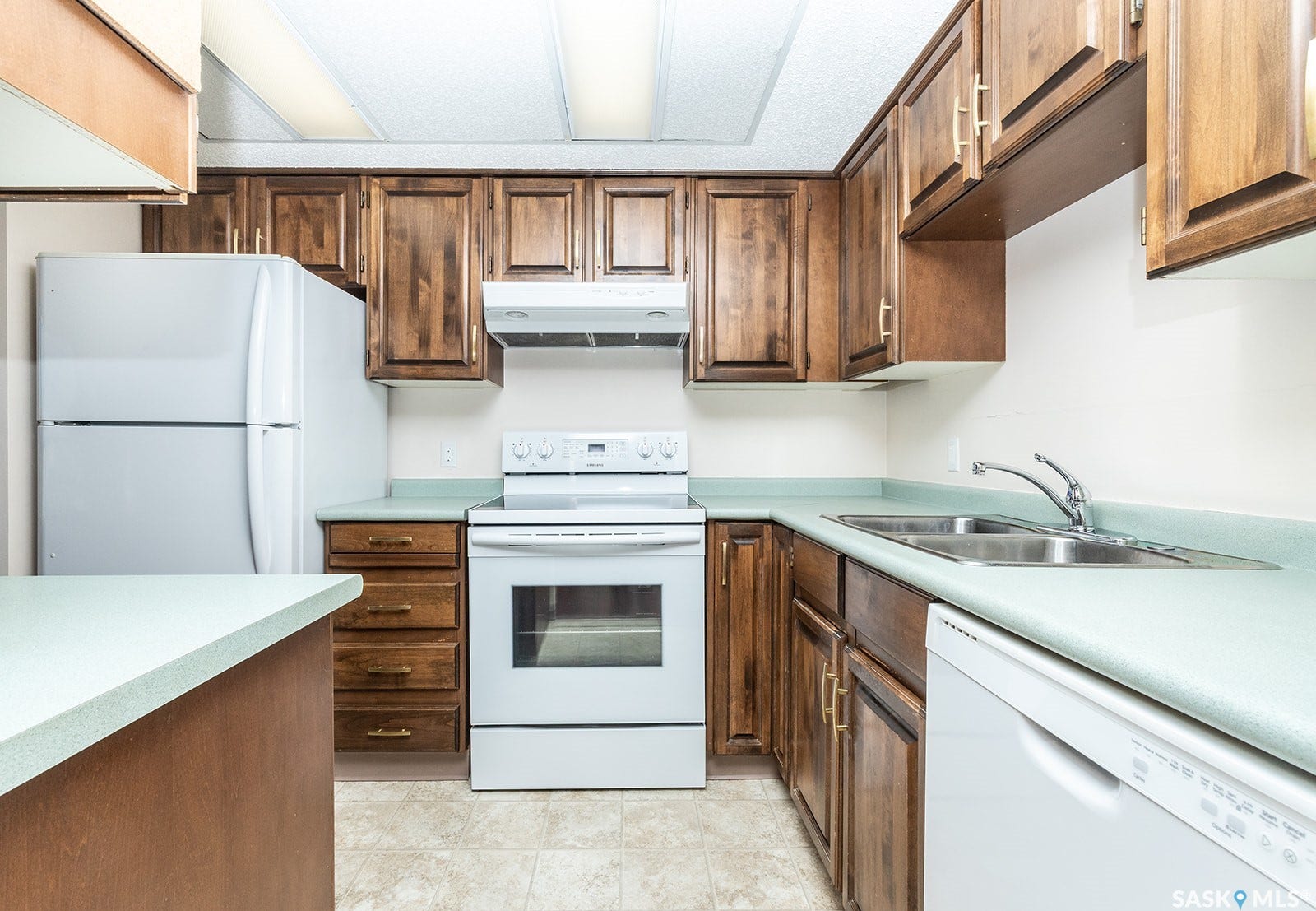 Kitchen featuring wood cabinetry, light-toned countertops, a double basin sink, white appliances and linoleum flooring
