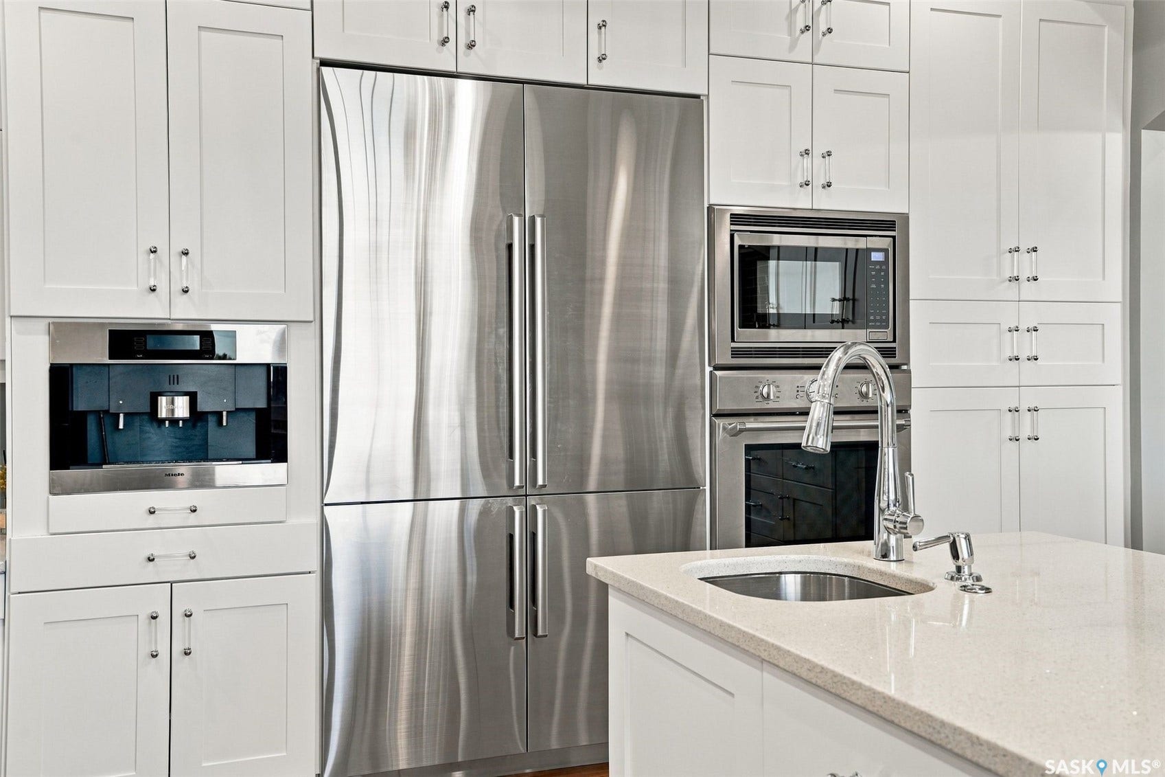 White shaker cabinetry with clear knob hardware, built-in coffee system, stainless steel refrigerator, built-in microwave, and oven