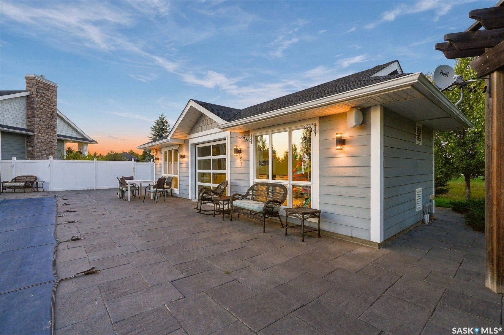 Expansive paver patio with a white privacy fence, an attached auxiliary structure featuring large windows, and wall-mounted exterior lighting