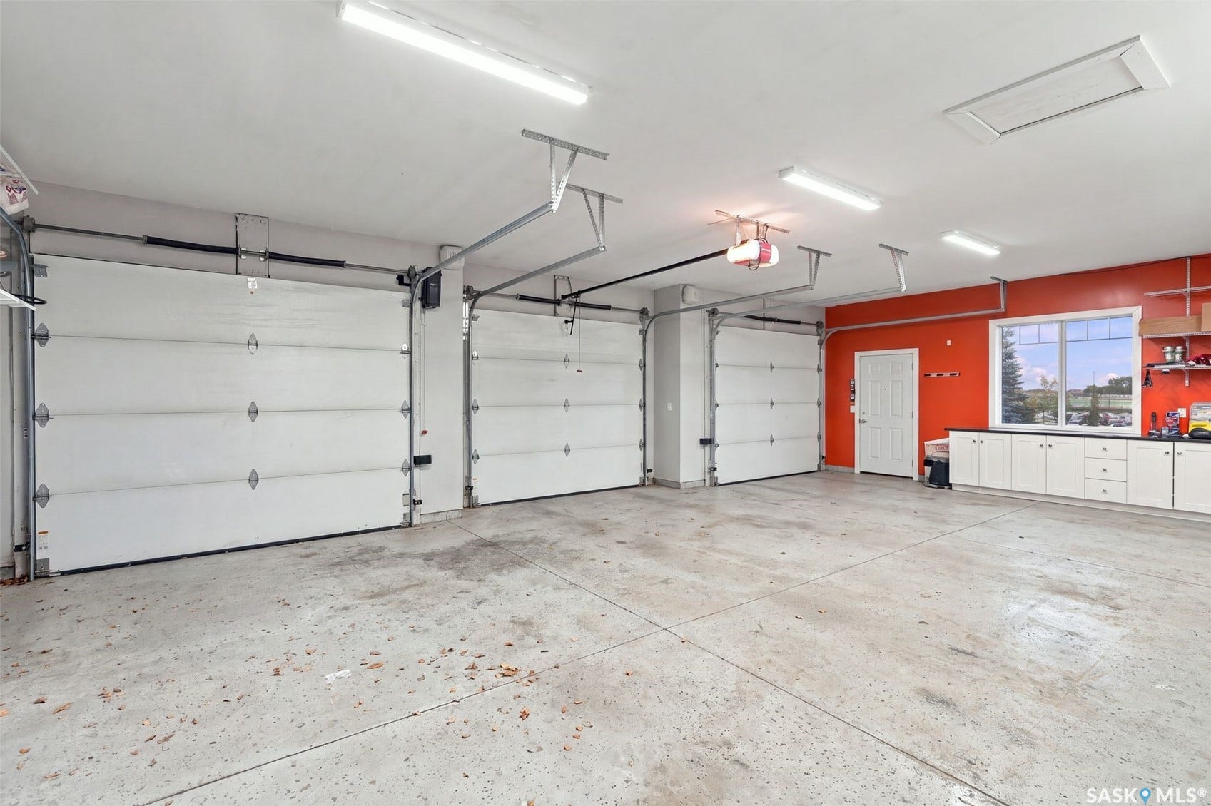 Three garage doors, concrete flooring, overhead lighting, white cabinetry, and a single window