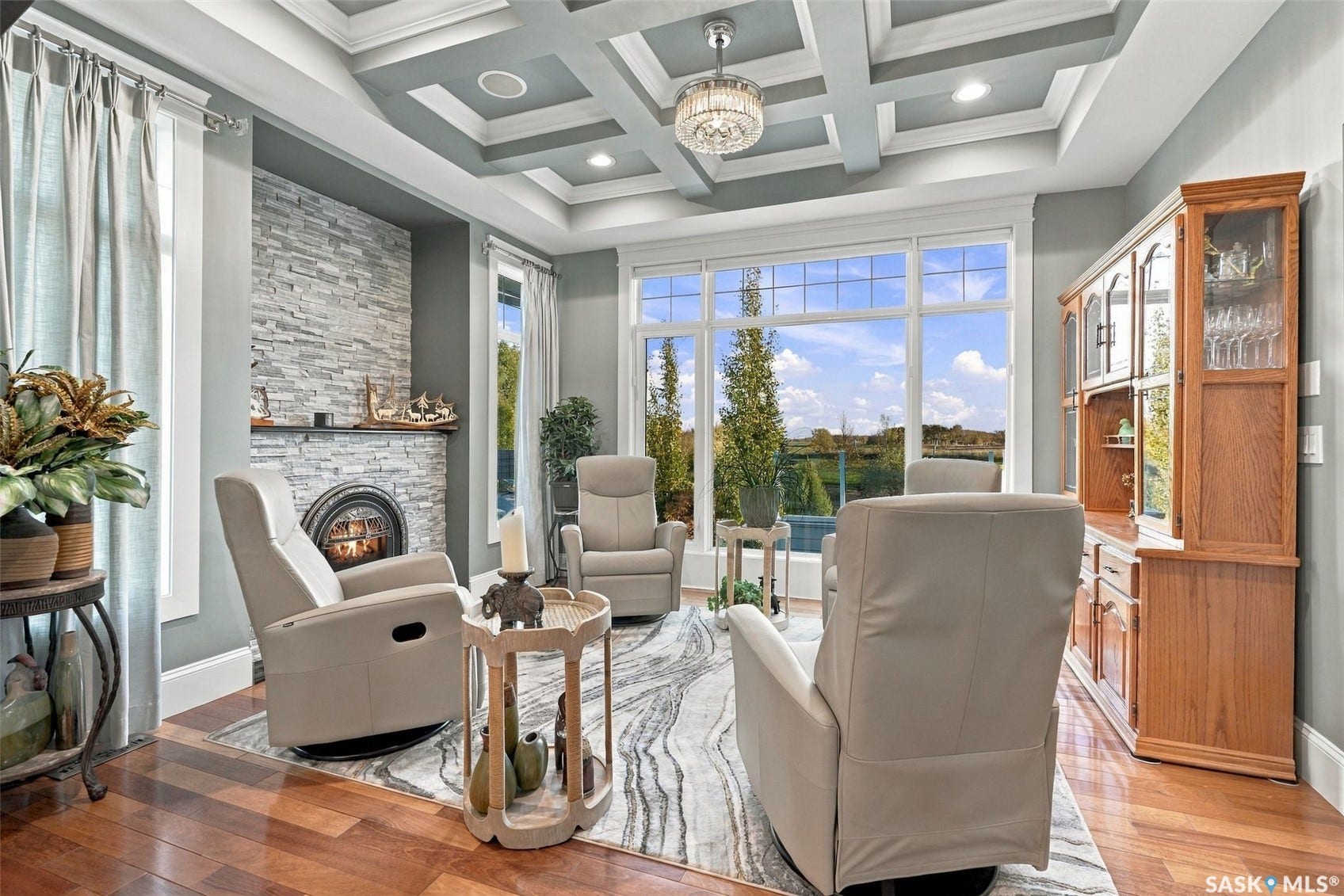 Coffered ceiling with a crystal chandelier, stacked stone fireplace, large picture windows, wood-finish flooring.