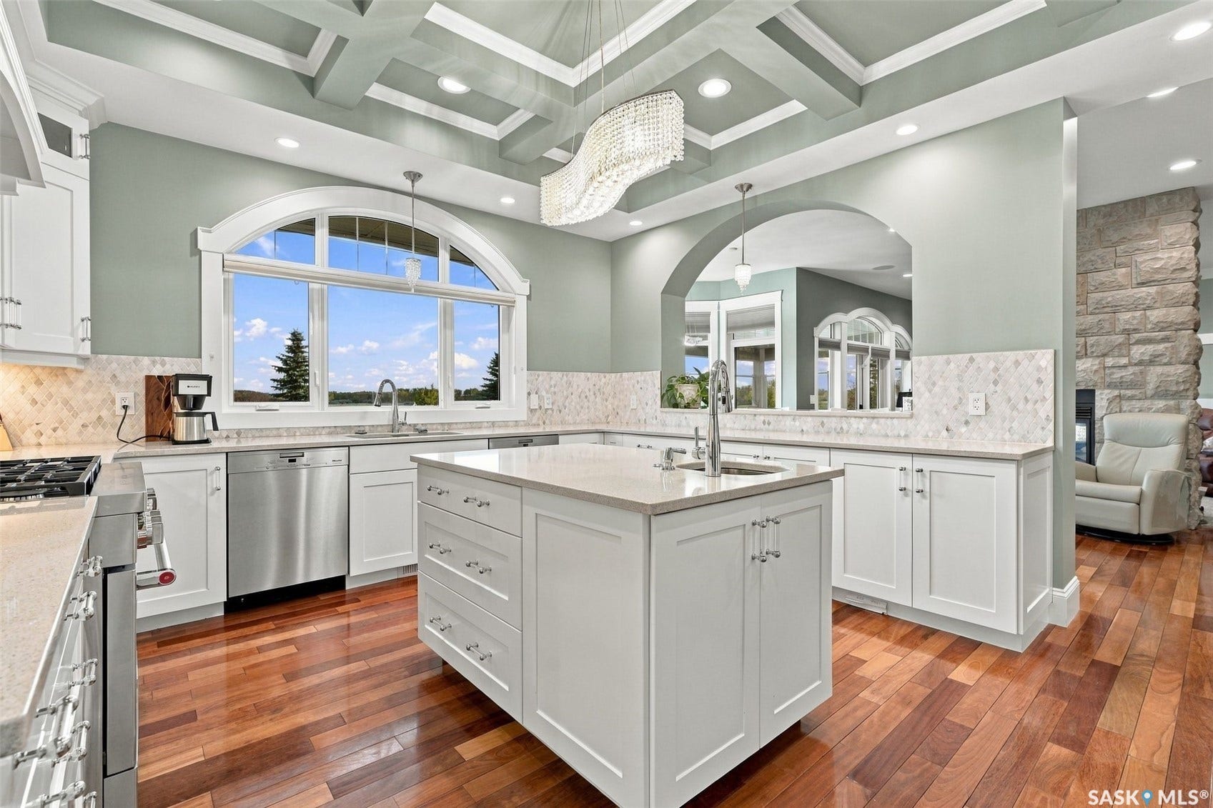 Kitchen featuring a multi-tier coffered ceiling, a large arched picture window, wood-finish flooring, a central island with an integrated sink, and white cabinetry with stone countertops