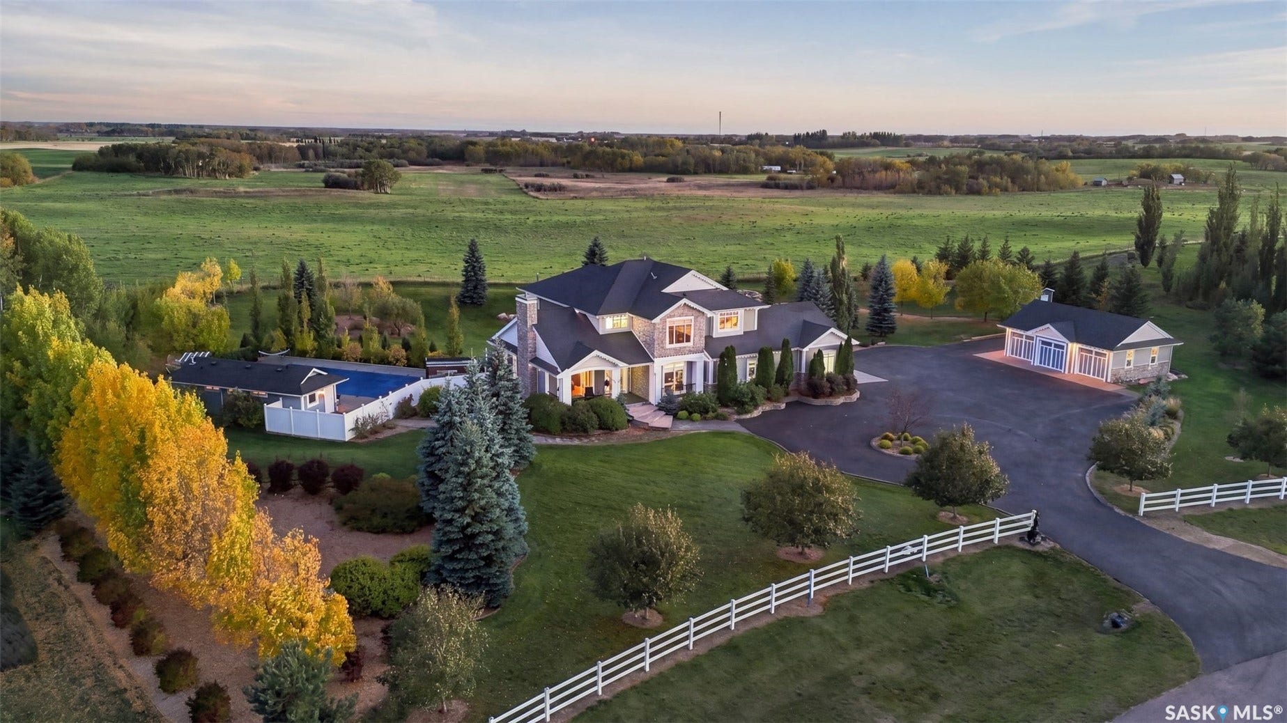 Expansive property featuring a grand residence, a separate detached garage structure, a fenced swimming pool with an accompanying pool house, a circular driveway, and white perimeter fencing