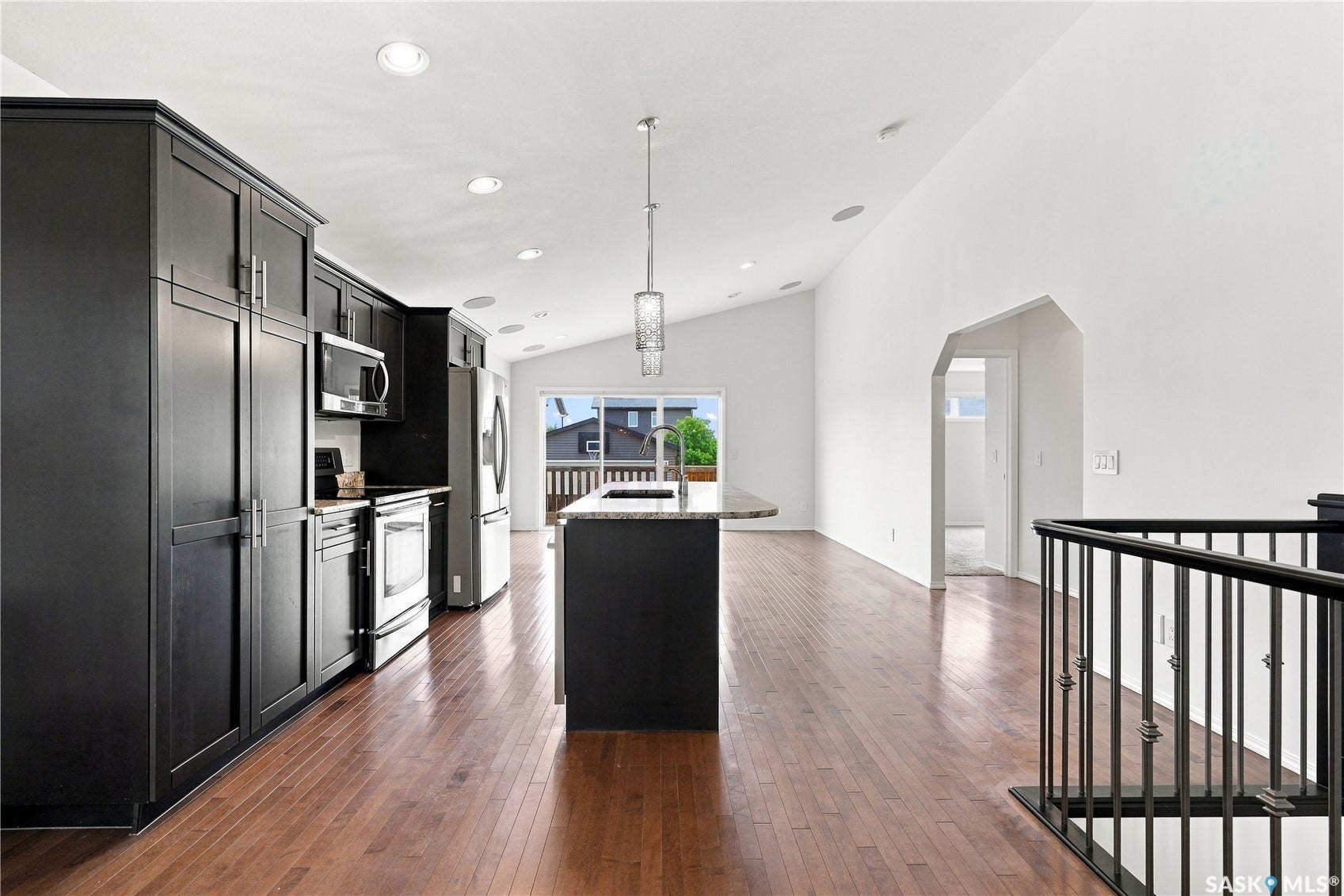 Vaulted ceiling with recessed lighting, wood-finish flooring, black cabinetry, stainless steel appliances, and a kitchen island with a sink