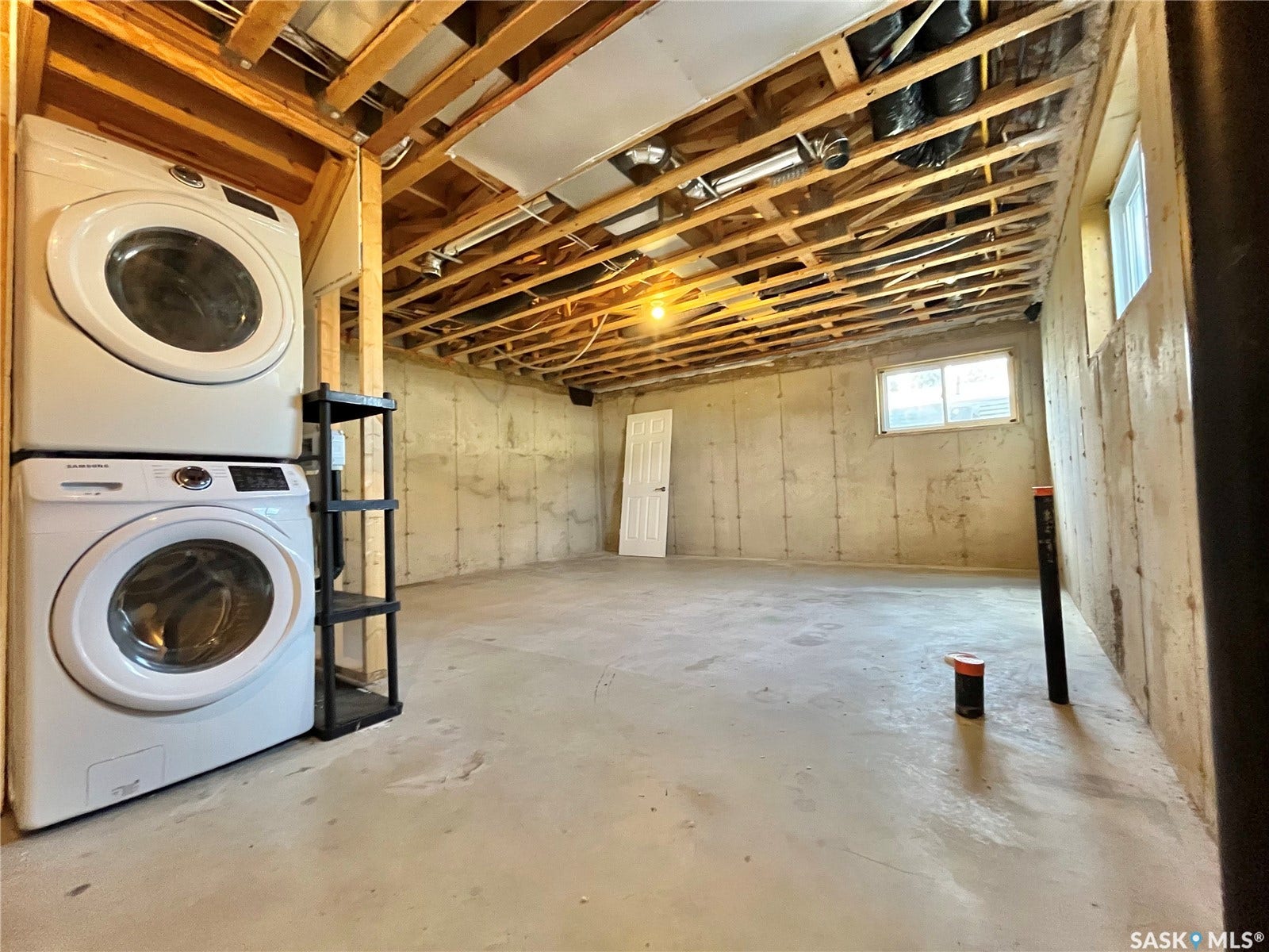 Unfinished basement with concrete flooring and exposed ceiling joists, featuring a stacked washer and dryer unit, a white interior door, and two windows