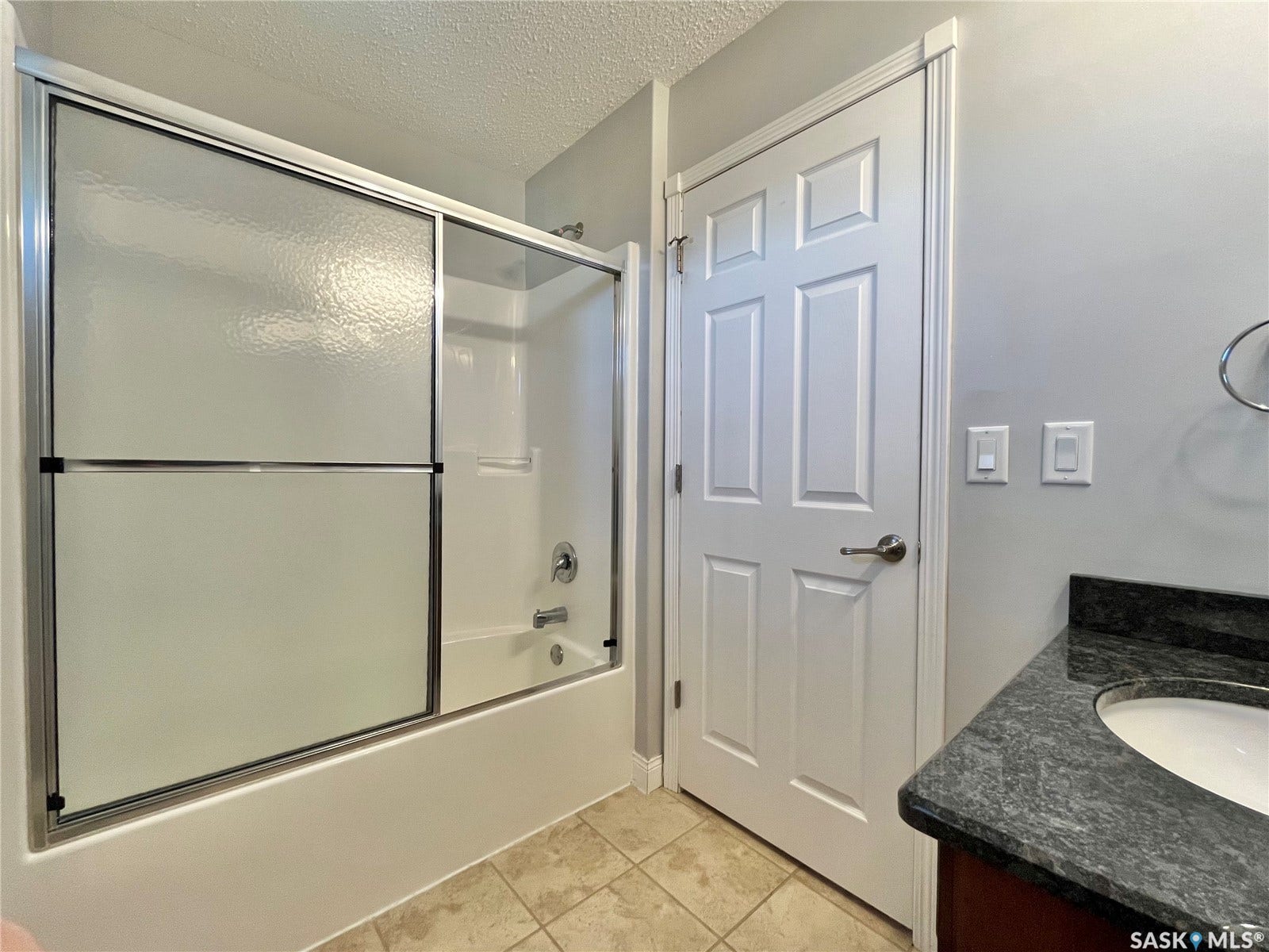 Built-in bathtub with sliding glass enclosure, vanity with dark stone countertop, undermount sink, and tiled flooring