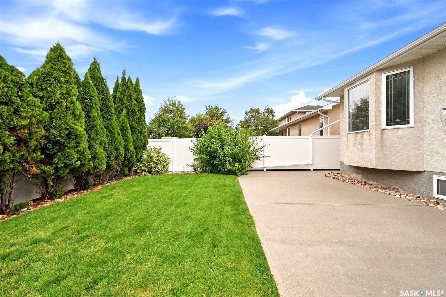 Expansive side yard featuring a lush lawn, mature evergreen privacy trees, a white panel fence, a concrete pathway, and a stucco exterior