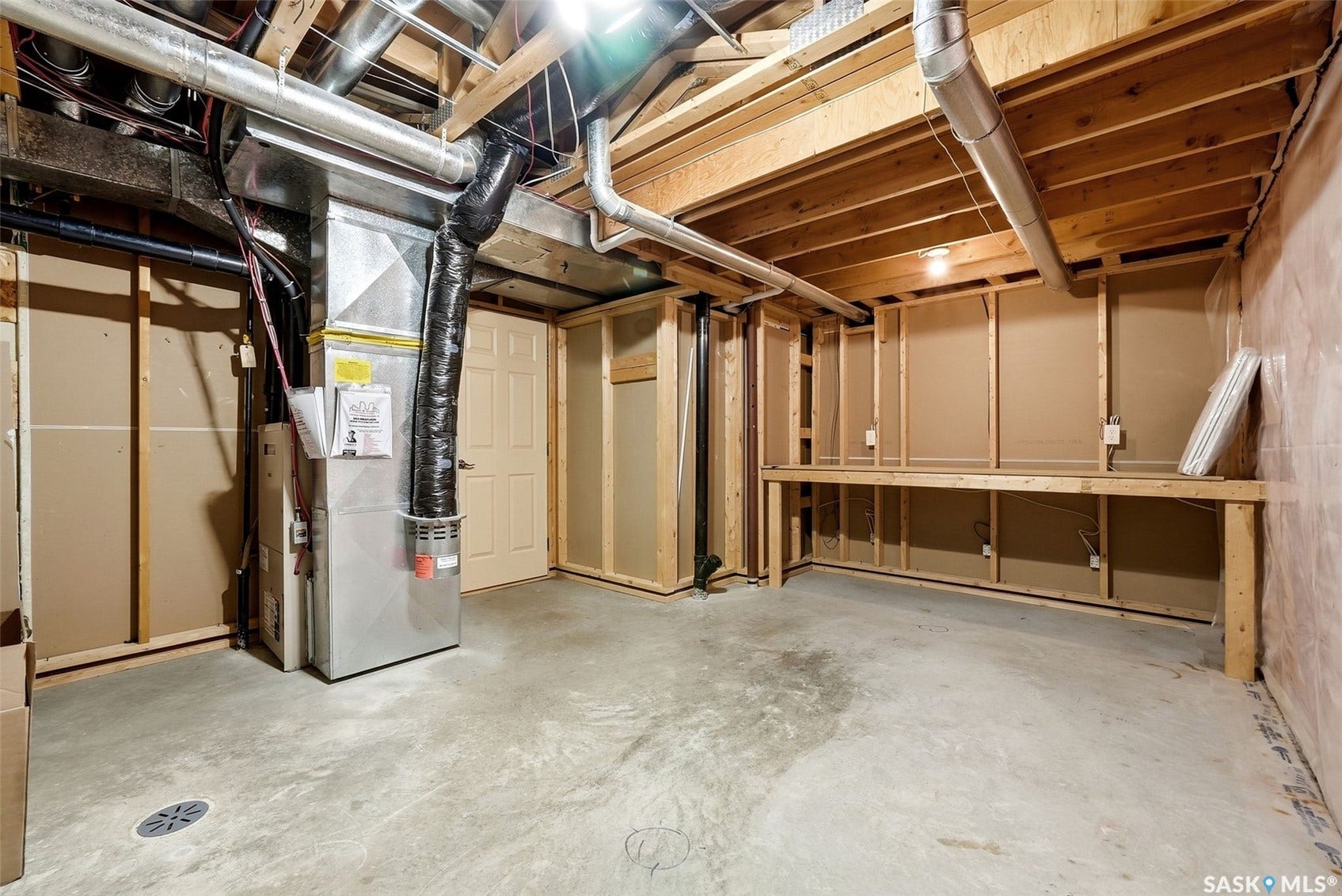 Unfinished basement with exposed ceiling joists, concrete flooring, a built-in workbench, and utility access