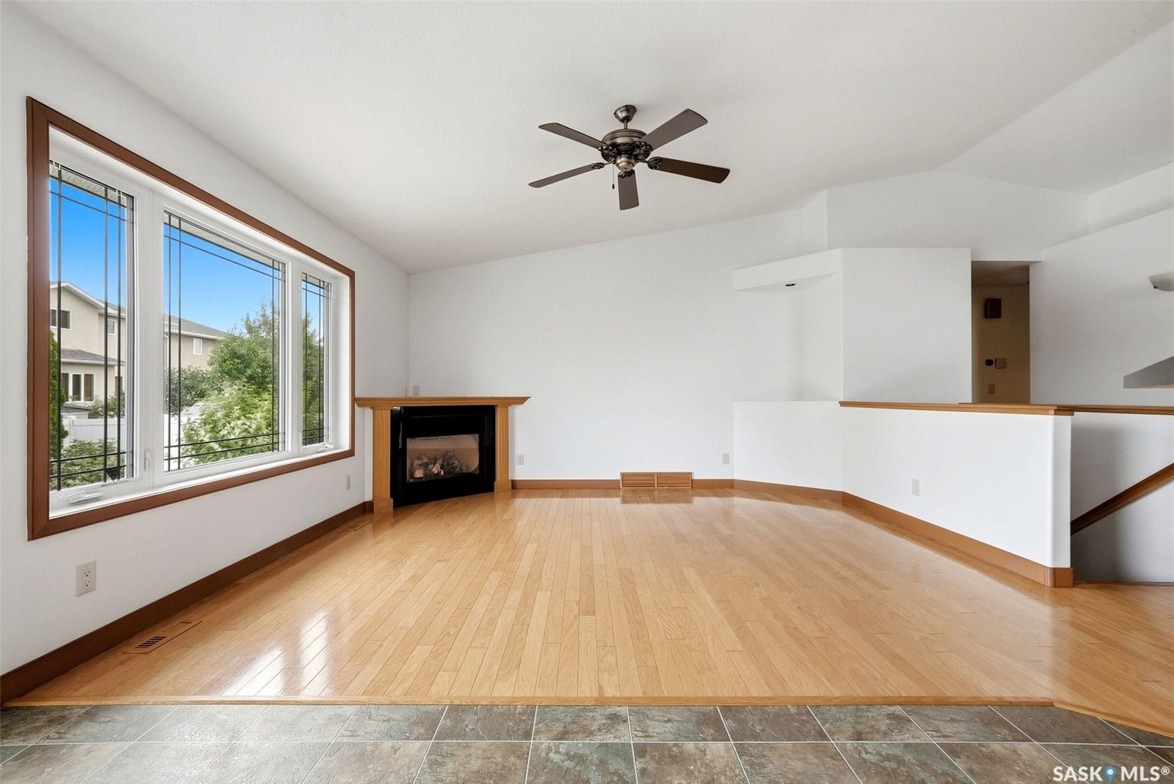 Hardwood flooring, corner fireplace with wood surround, large window with mullions, vaulted ceiling, and a ceiling fan