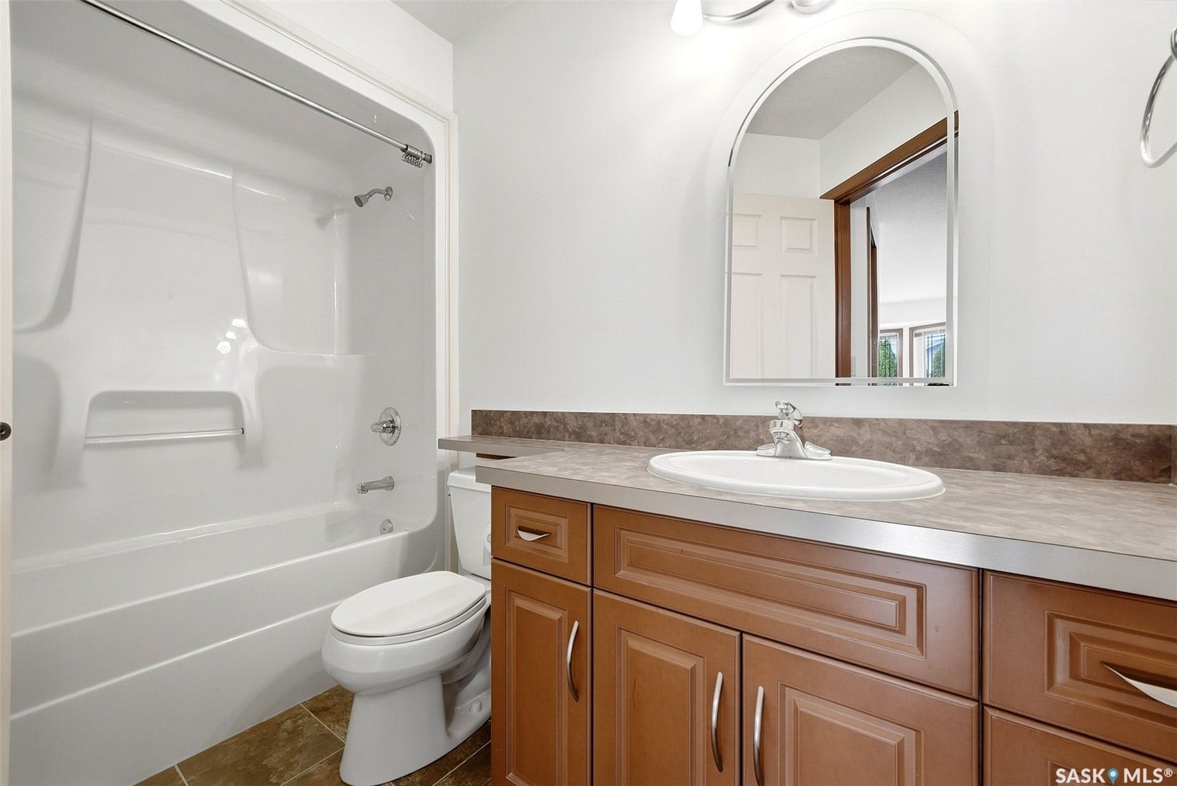 White shower/tub combination, single vanity with wood-finish cabinetry, countertop, and an arched mirror