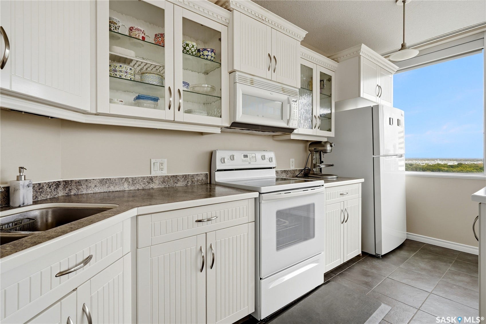 1914 Hamilton Street, #1007, Regina SK S4P 3N6 White shaker-style cabinetry with glass-front accents, dark stone-finish countertops, stainless steel sink, and white appliances, all complemented by dark floor tiling