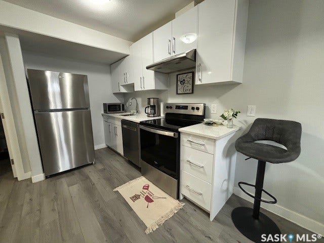 Stainless steel appliances, wood-finish flooring, white flat-panel cabinetry, white solid surface countertops, and an undermount sink