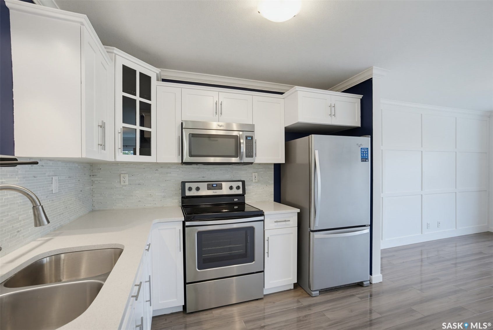 White cabinetry with brushed nickel hardware, stainless steel appliances, light-toned countertop, mosaic tile backsplash, and wood-finish flooring