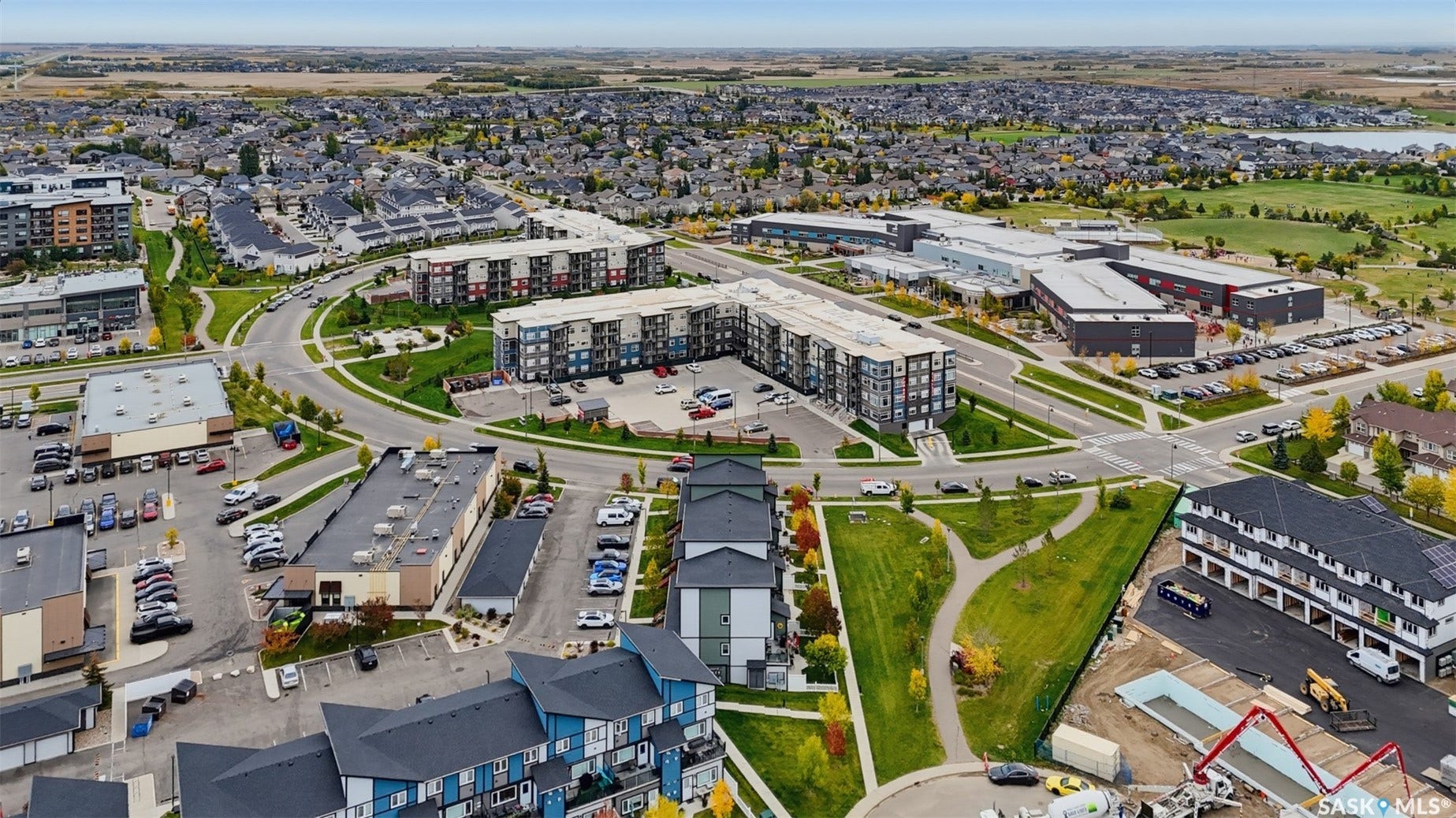 Aerial perspective showcasing a residential and commercial development, featuring multiple multi-story buildings, extensive paved parking areas, and landscaped green spaces with established pathways