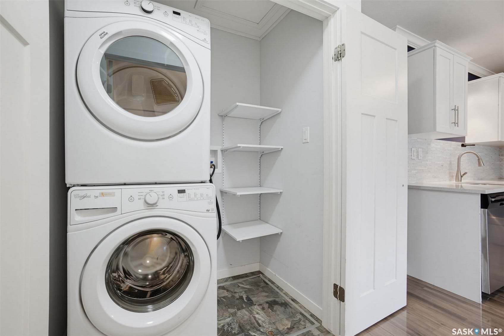 Stackable washer and dryer, built-in shelving, gray tile flooring, white shaker cabinets, and stainless steel kitchen appliances
