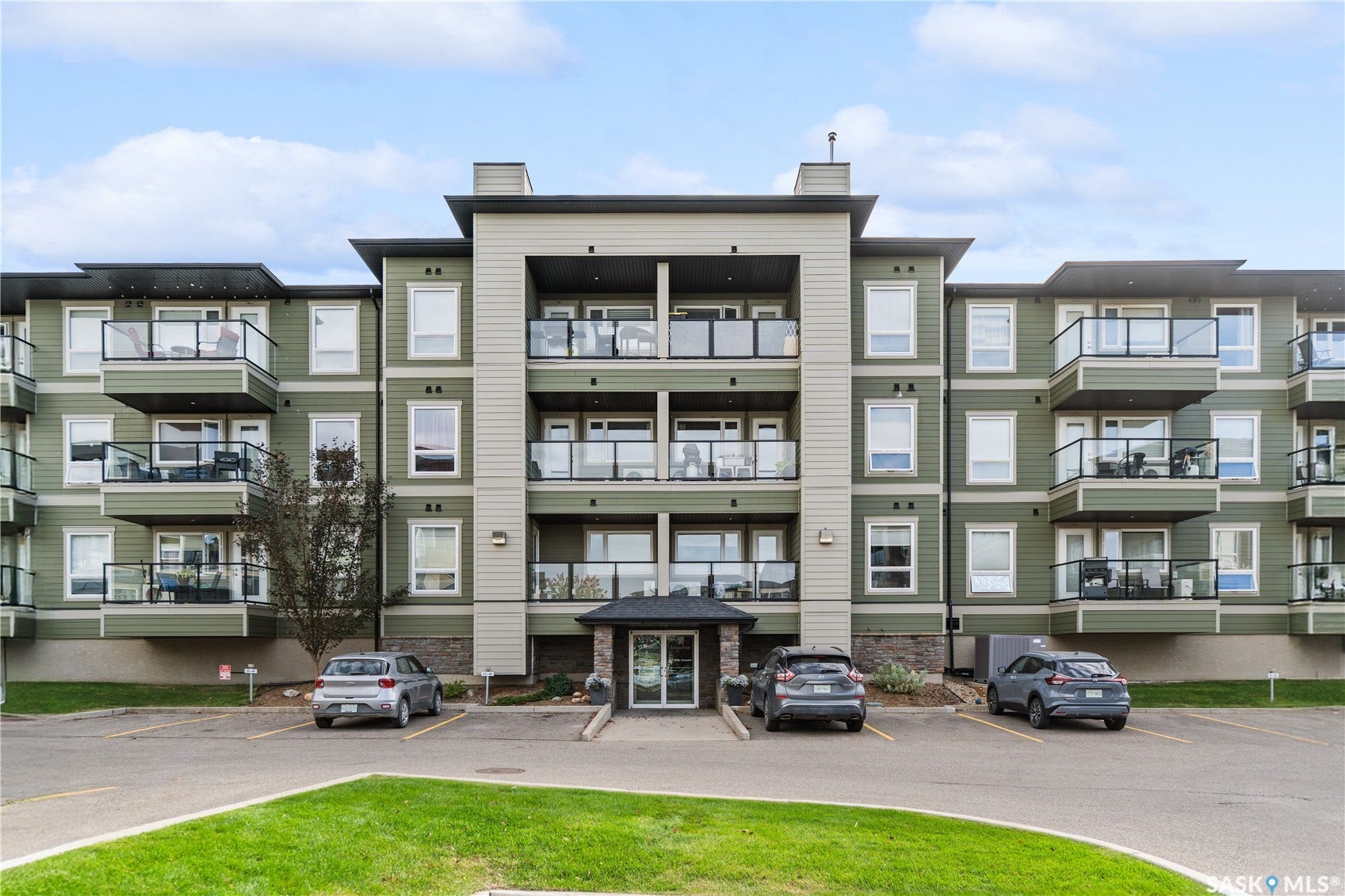 Multi-story building featuring private balconies with glass railings, a stone-clad entrance, ample paved parking, and a manicured green space