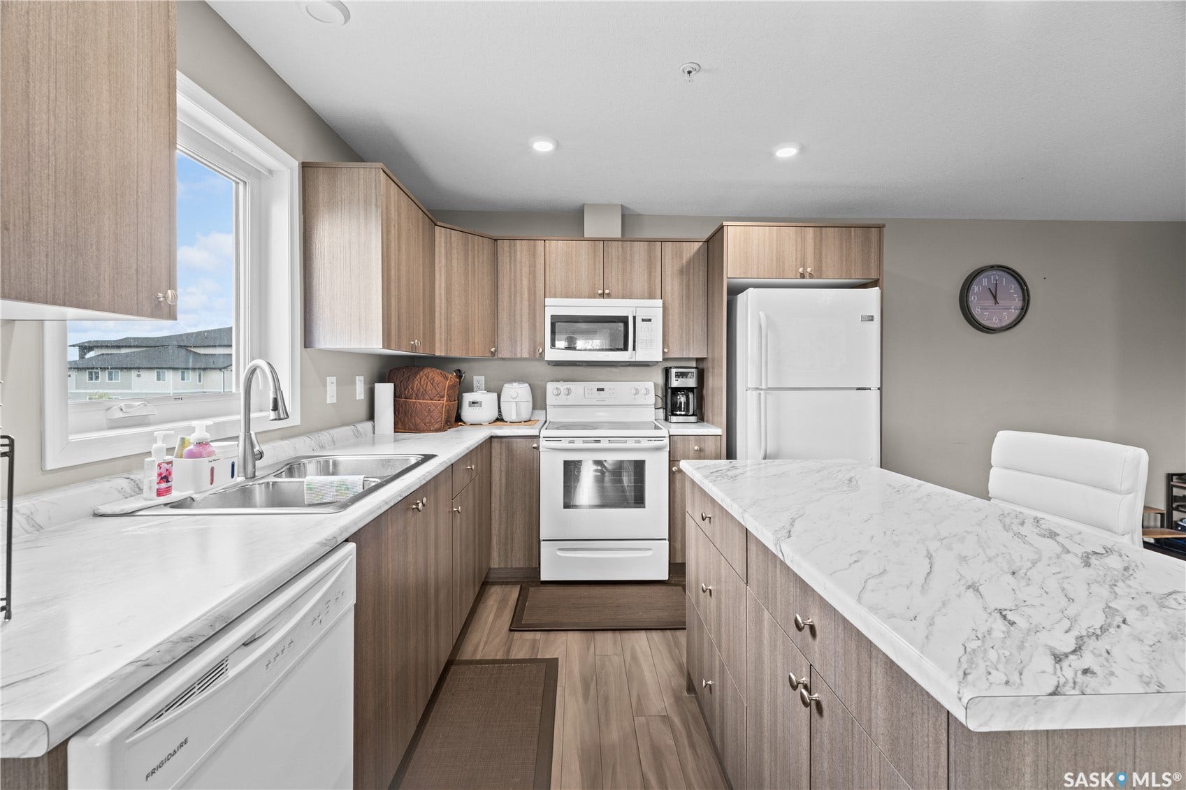 Kitchen featuring wood-finish cabinetry, white appliances, a double basin stainless steel sink, and light-toned countertops with a central island