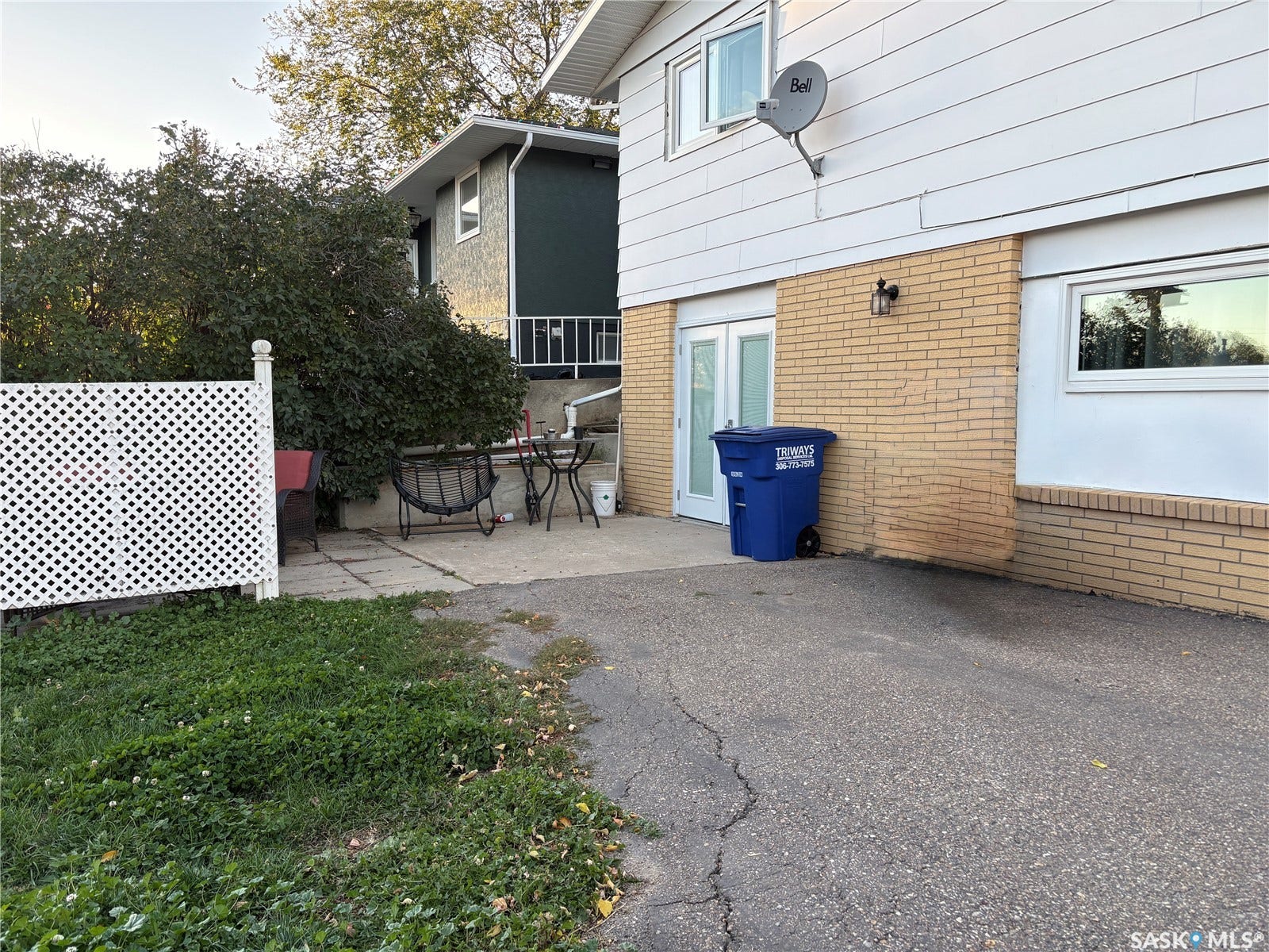 Paved driveway leading to a concrete patio with white lattice fencing and mature landscaping