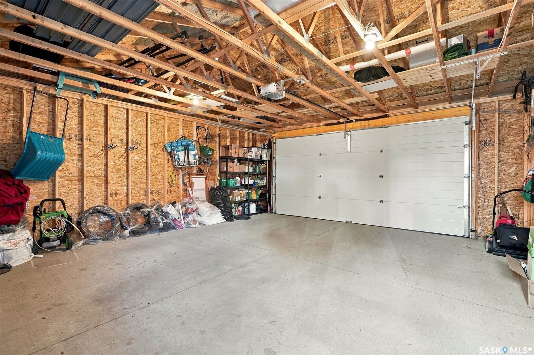 919 Kolynchuk Bend, Saskatoon SK S7T 0V9 Spacious garage featuring exposed wood framing, concrete flooring, overhead wood shelving, and a segmented garage door