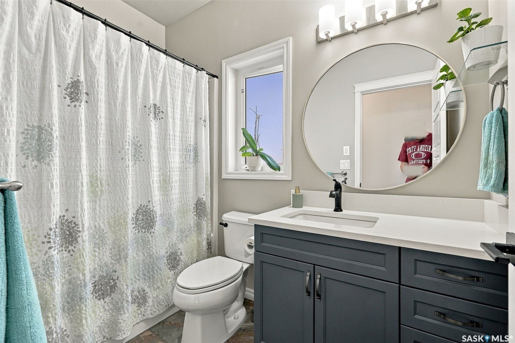 919 Kolynchuk Bend, Saskatoon SK S7T 0V9 Vanity with dark cabinetry and white countertop, integrated sink, large round mirror, and wall-mounted light fixture