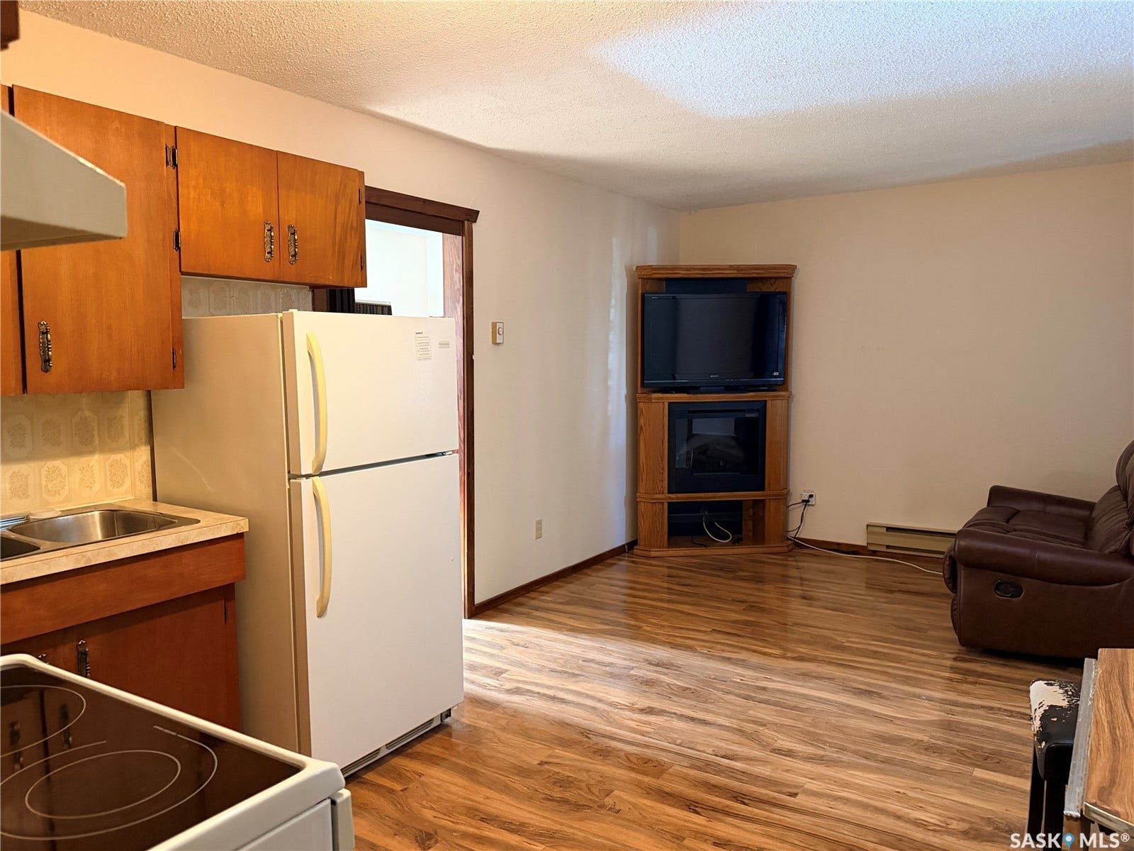 Open-concept living space with wood-finish flooring, a built-in corner entertainment unit, and a kitchen area featuring wooden cabinetry