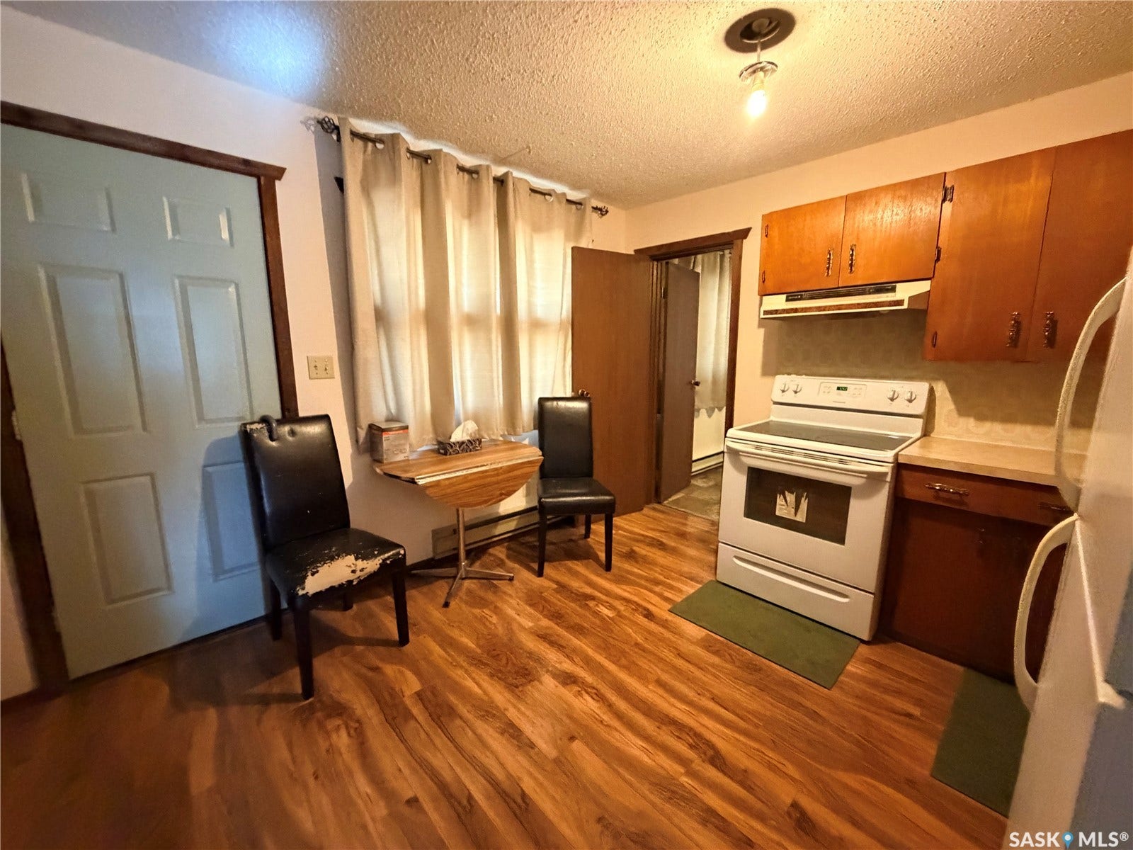 Wood-finish flooring throughout, white paneled interior door with dark wood trim, window with privacy curtains, built-in wooden cabinetry, and a freestanding electric range