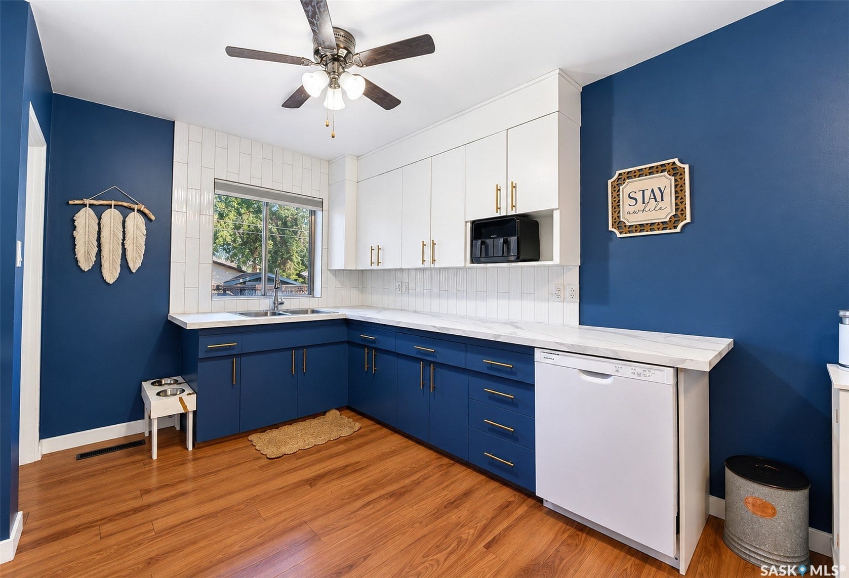 Well-appointed kitchen featuring wood-finish flooring, white upper cabinetry, blue lower cabinetry, white subway tile backsplash, and a ceiling fan with integrated lighting