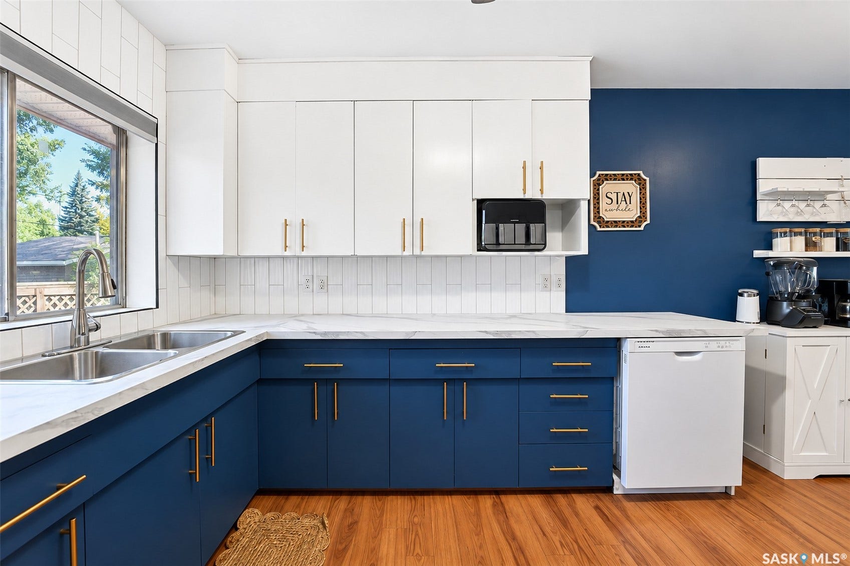 Contemporary kitchen featuring two-tone cabinetry with white upper cabinets and deep blue lower cabinets accented by brushed gold hardware, white subway tile backsplash, quartz countertops, stainless steel appliances including a built-in coffee maker, and warm wood-finish flooring throughout