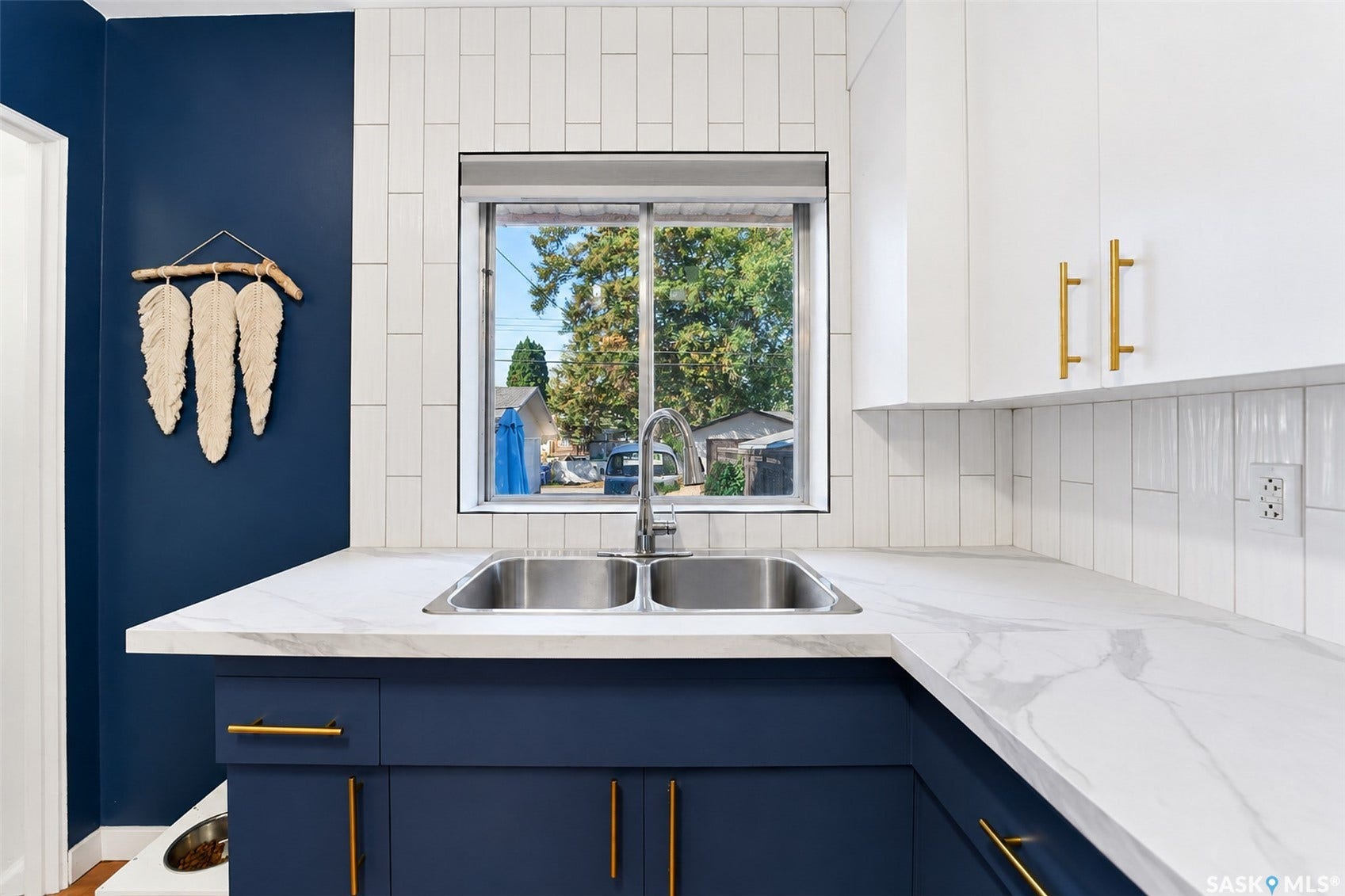 Crisp white slab cabinetry with brass hardware, a dual basin stainless steel sink, white marble-look countertops, and a white vertical subway tile backsplash