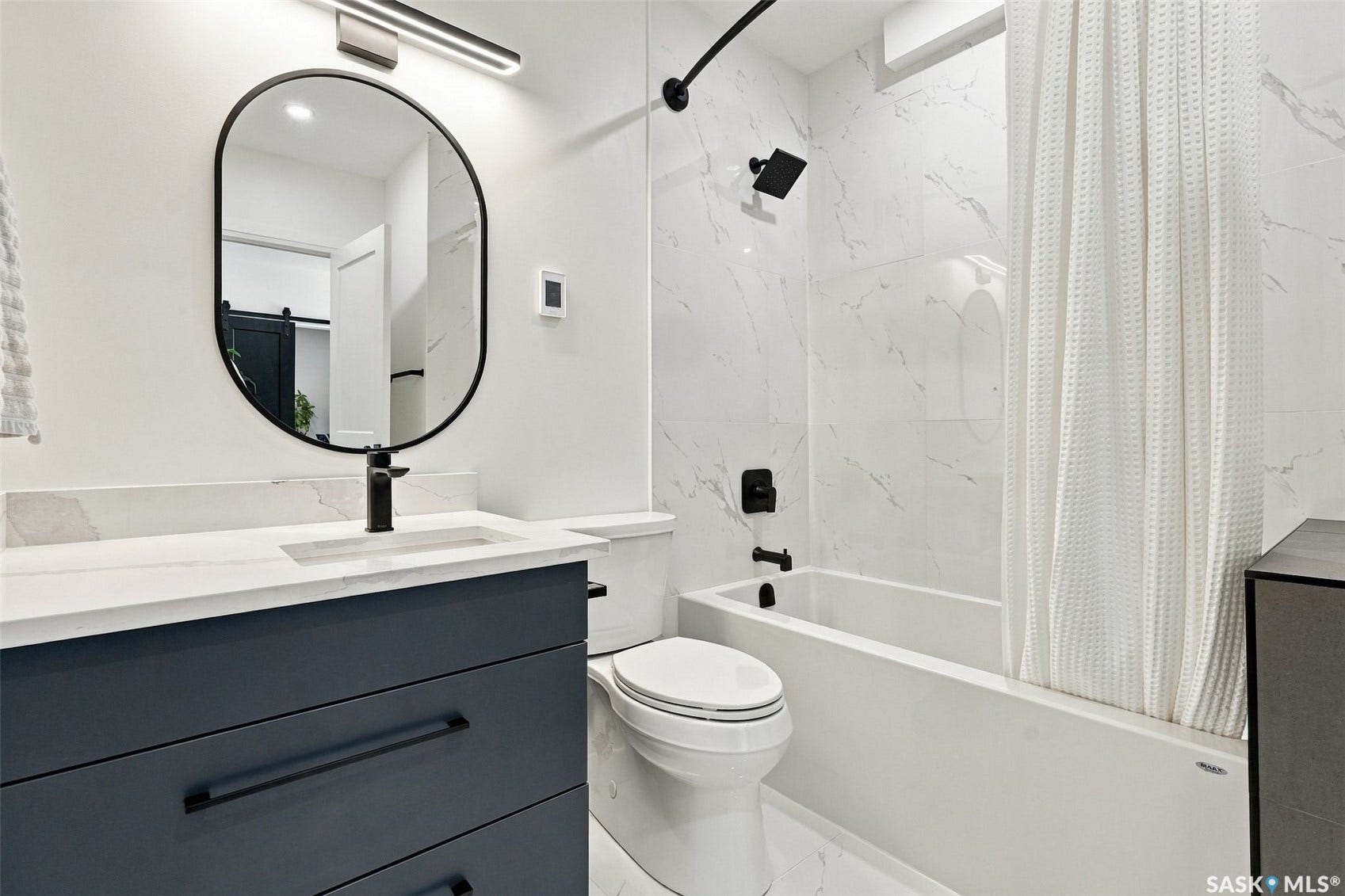 6221 Koep Avenue, Regina SK S4X 0J2 Modern bathroom featuring a matte black framed oval mirror, dark gray vanity with black hardware, white countertop with integrated sink, white tiled shower surround, and a white bathtub with black fixtures