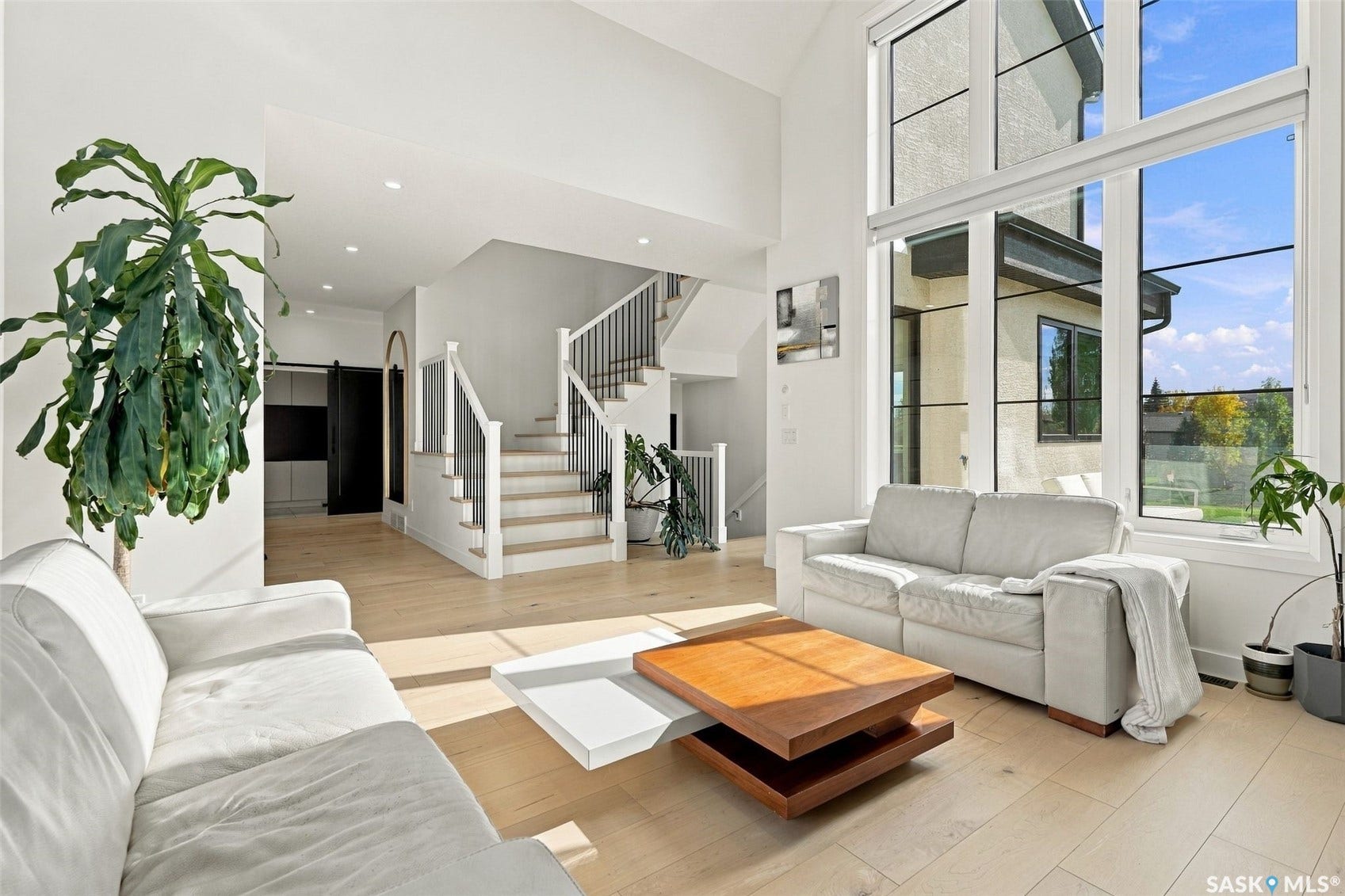 6221 Koep Avenue, Regina SK S4X 0J2 Double-height ceilings, expansive window wall with black trim, light wood-finish flooring, staircase with white risers and dark metal balusters, and recessed ceiling lighting