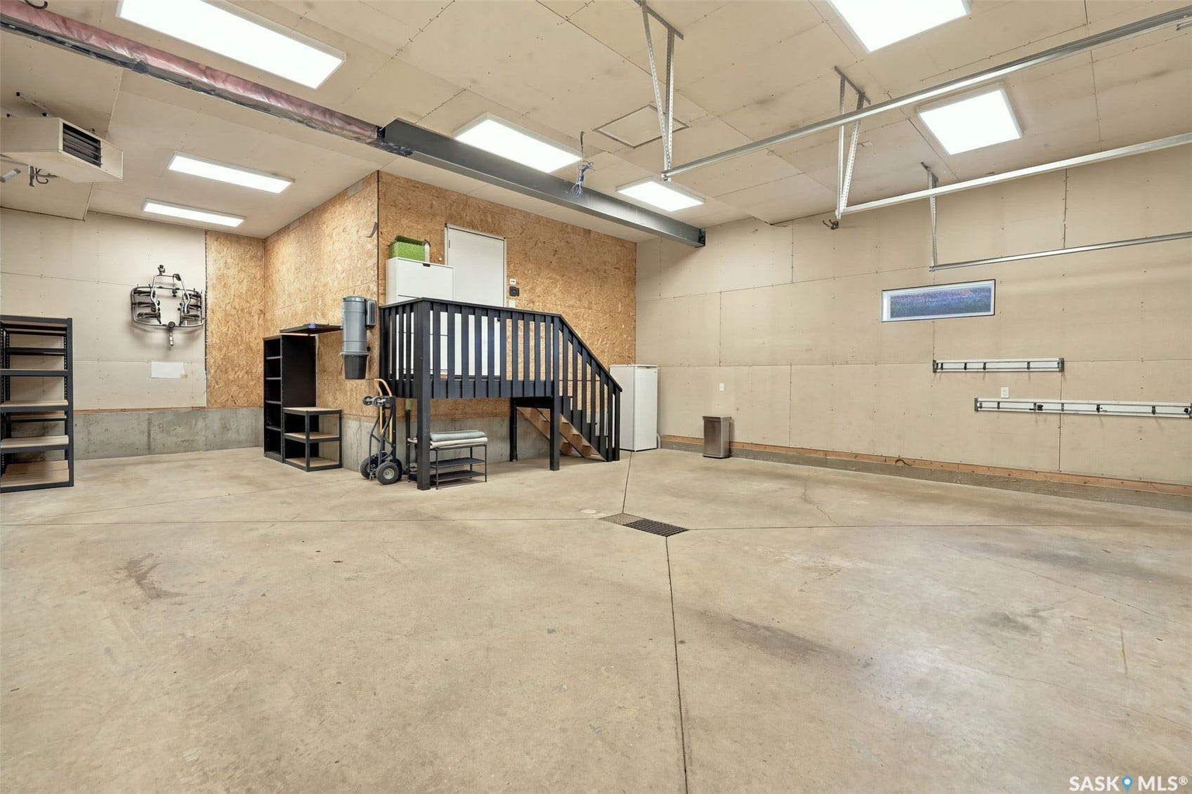 6221 Koep Avenue, Regina SK S4X 0J2 Spacious utility area featuring a concrete floor, built-in staircase with railing, overhead lighting fixtures, and a wall-mounted heater