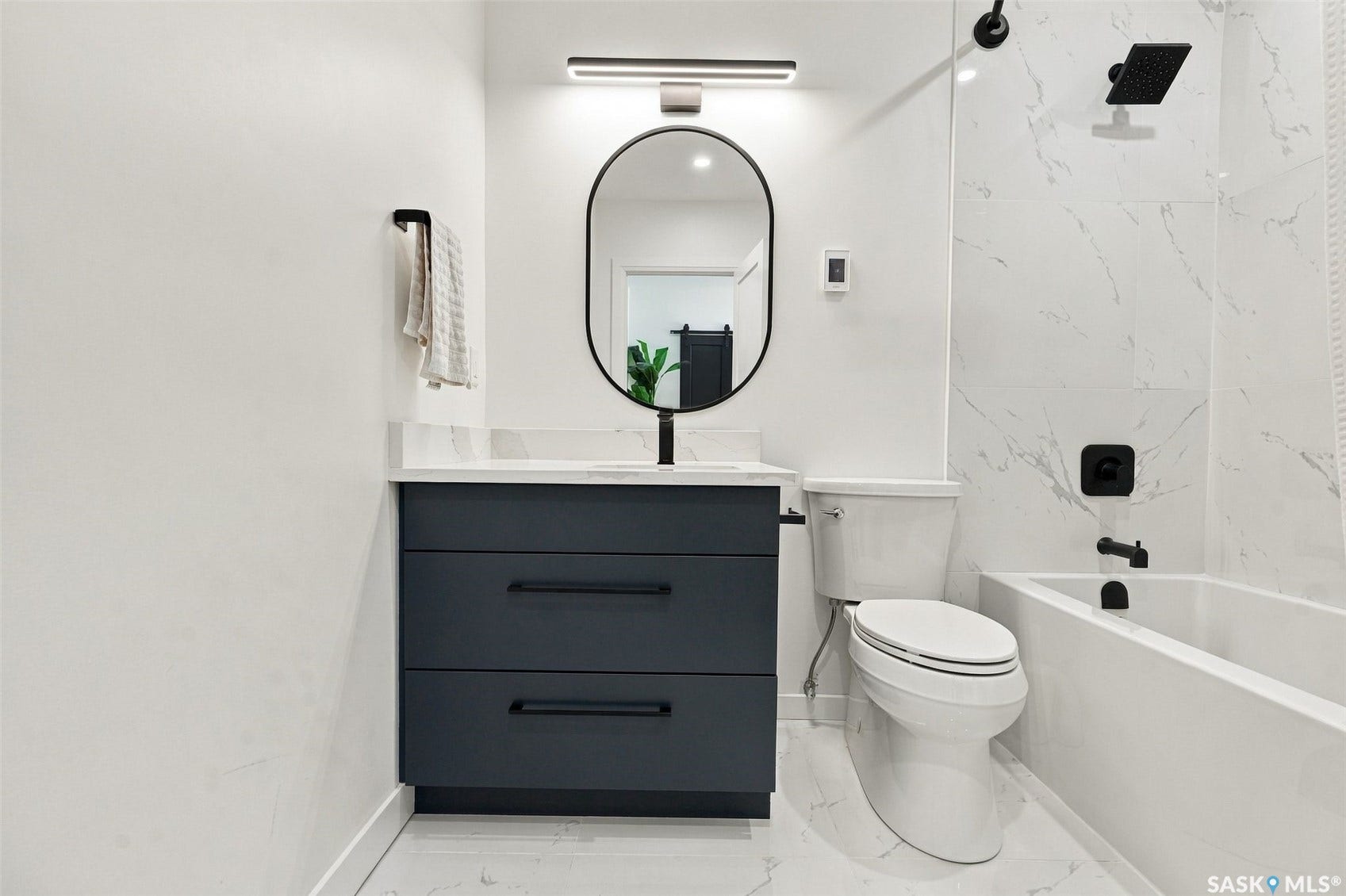 6221 Koep Avenue, Regina SK S4X 0J2 Modern bathroom featuring a dark vanity with a white countertop, oval mirror, wall-mounted light fixture, and a white tub with subway tile surround