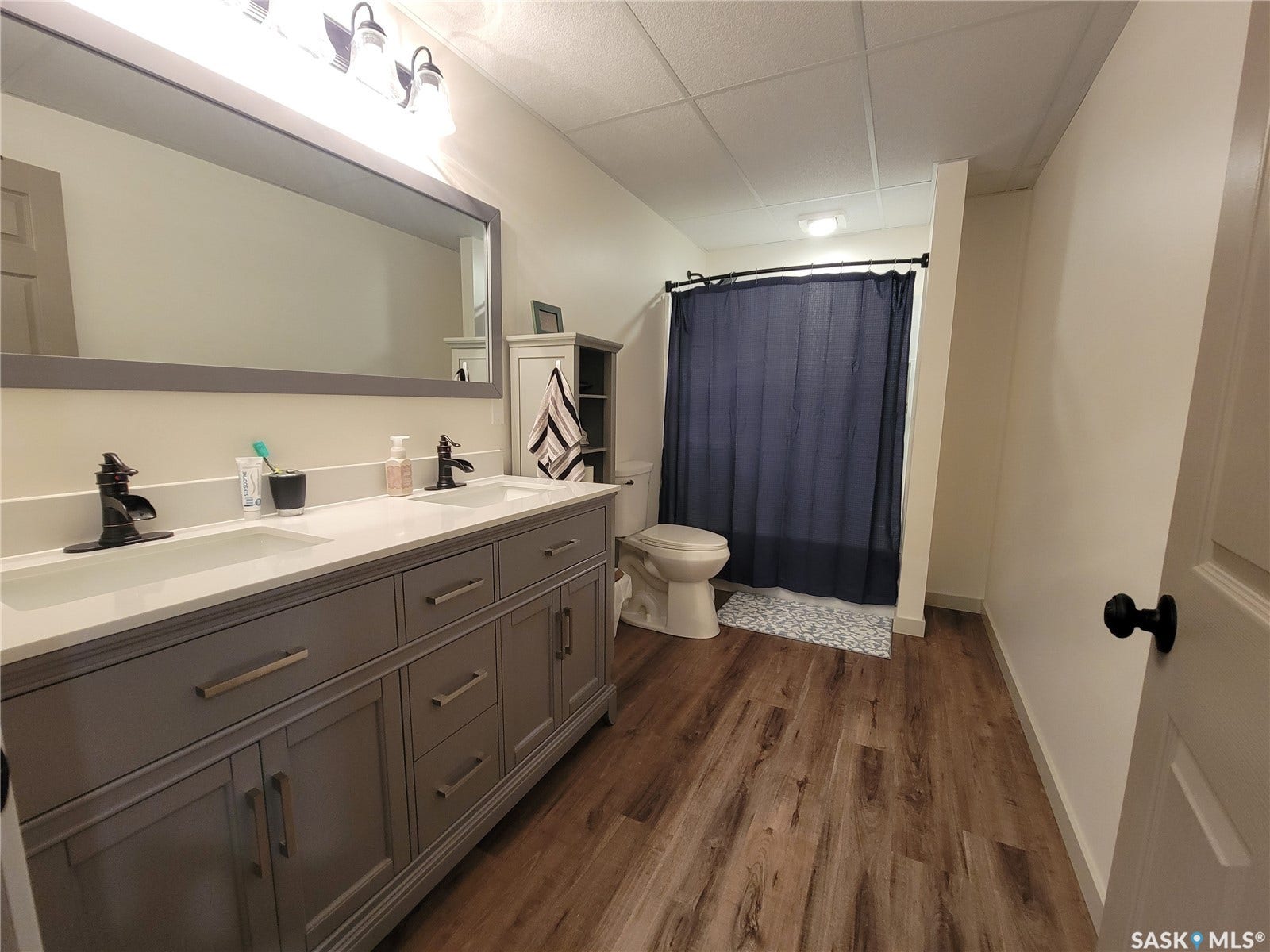Double vanity with white countertops, wood-finish flooring, framed mirror with integrated lighting, and a built-in shower area
