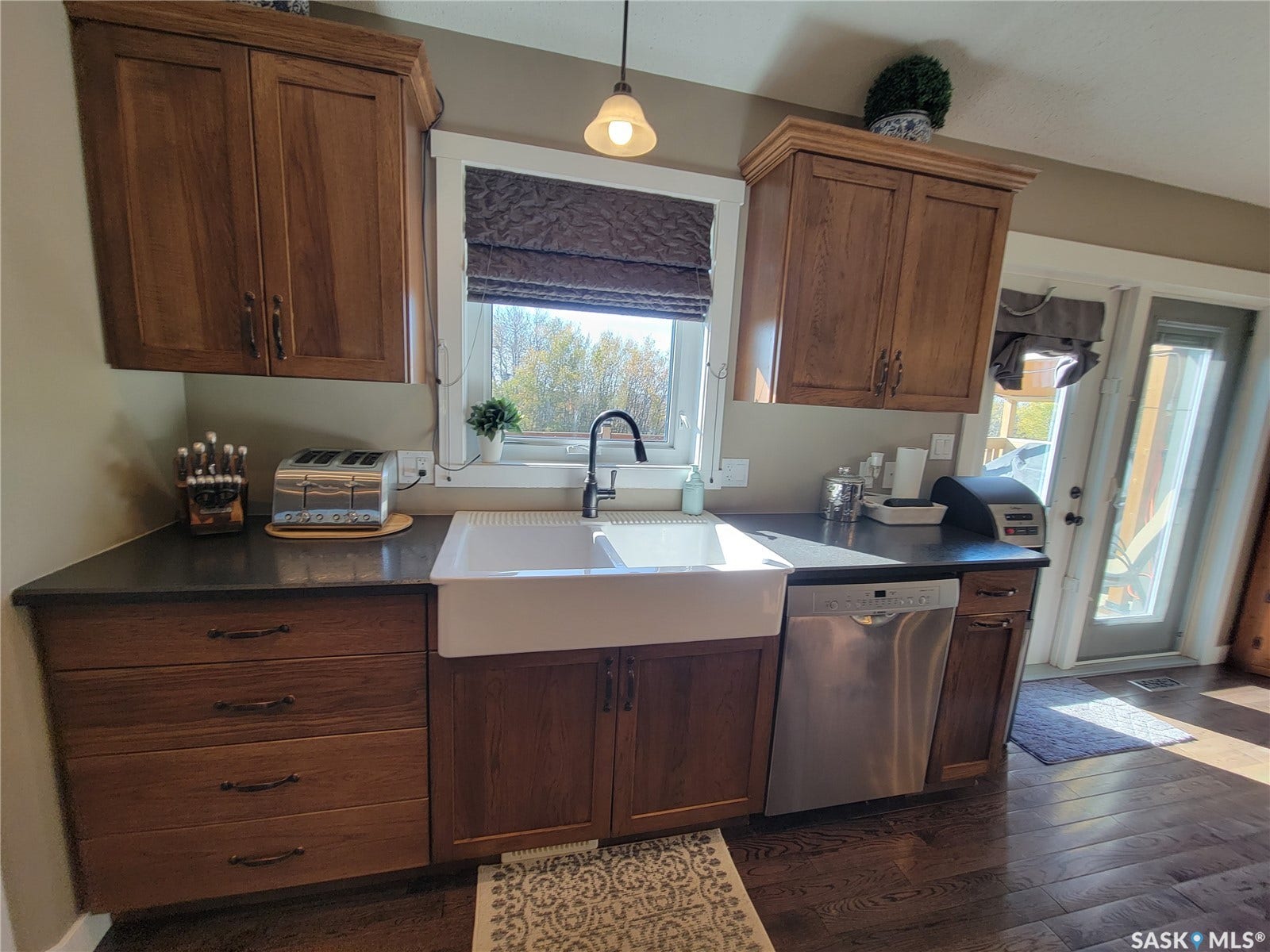 Wood-finish cabinetry, solid surface countertops, farmhouse-style sink with a matte black faucet, stainless steel dishwasher, and dark wood-finish flooring