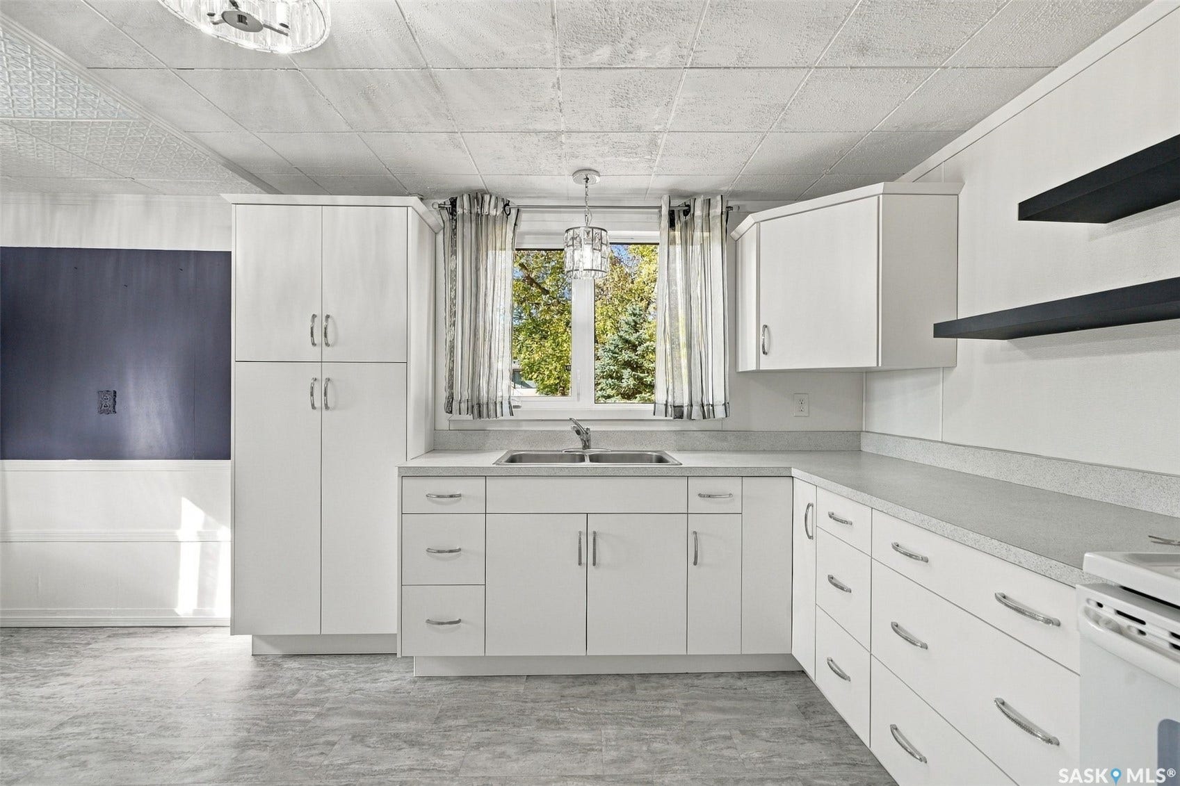 White cabinetry with stainless steel hardware, a dual-basin sink, light-toned countertops, and floating wall shelves
