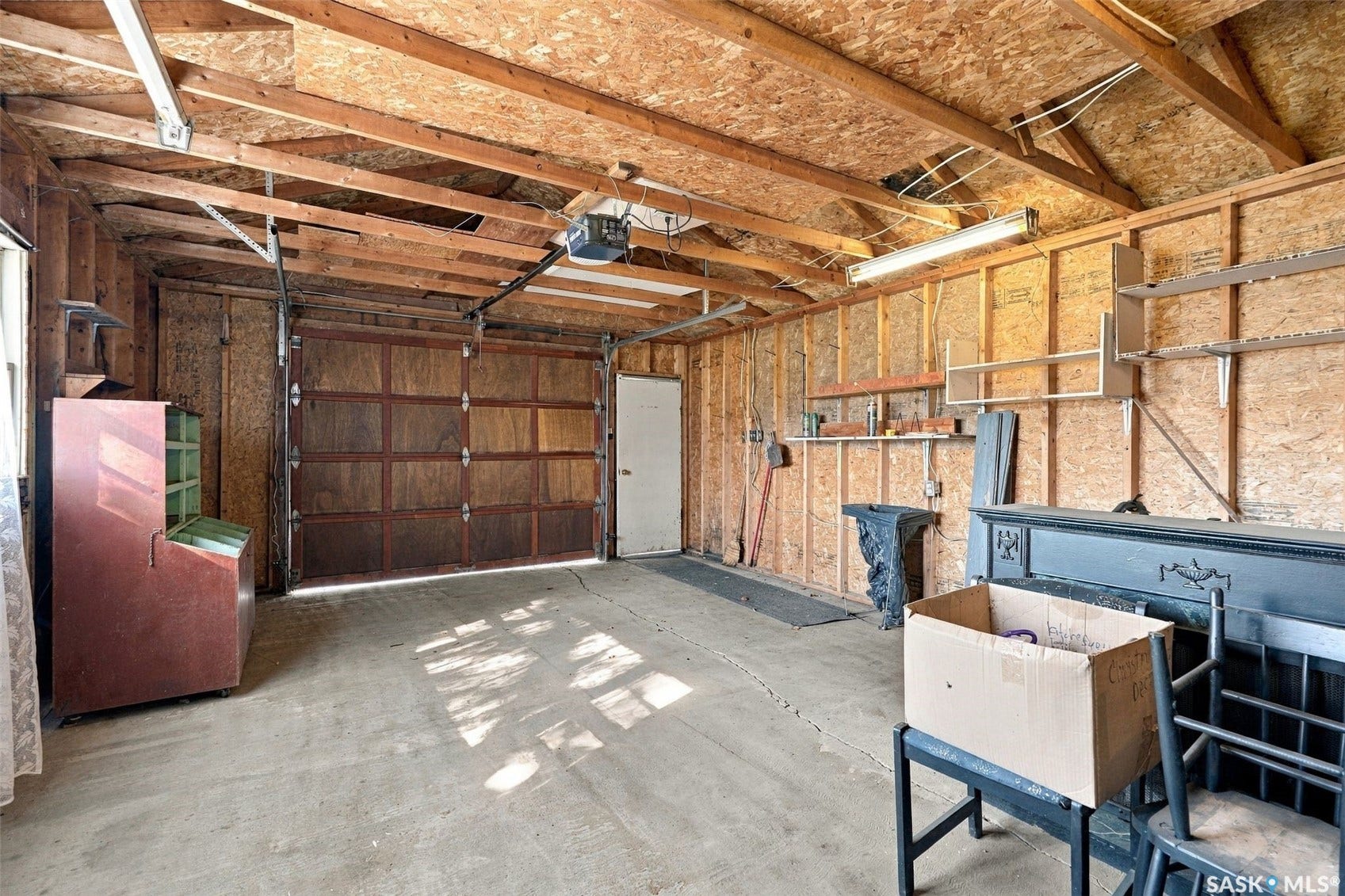 Concrete flooring, exposed wood beam ceiling, built-in shelving, overhead lighting, and a paneled garage door