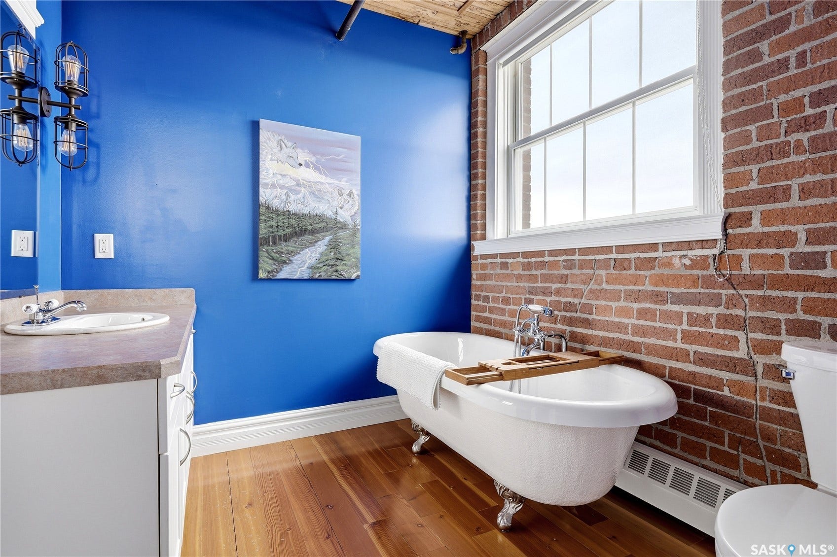 Wood-finish flooring, exposed brick accent wall, freestanding clawfoot tub, white window frame, and vanity with integrated sink