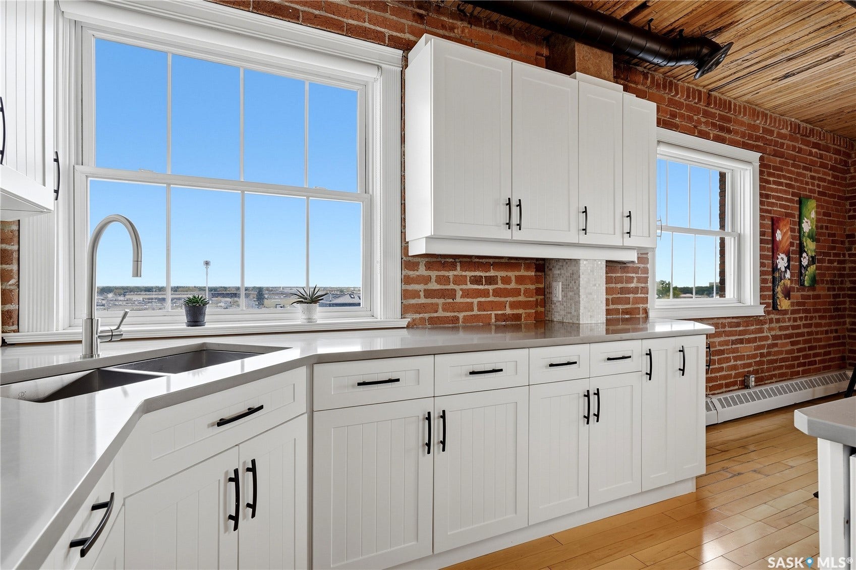 White shaker-style cabinetry with matte black hardware, stainless steel undermount sink with gooseneck faucet, light-toned countertop, exposed brick wall, and wood-finish flooring