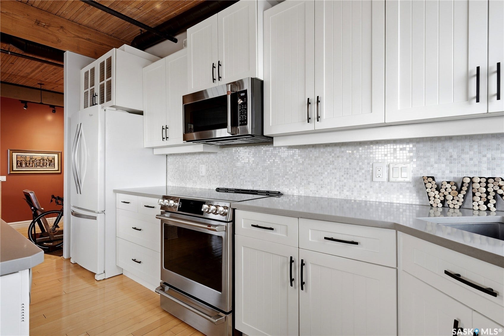 White shaker-style cabinetry with black hardware, stainless steel appliances, light-toned countertops, mosaic tile backsplash, and light wood-finish flooring