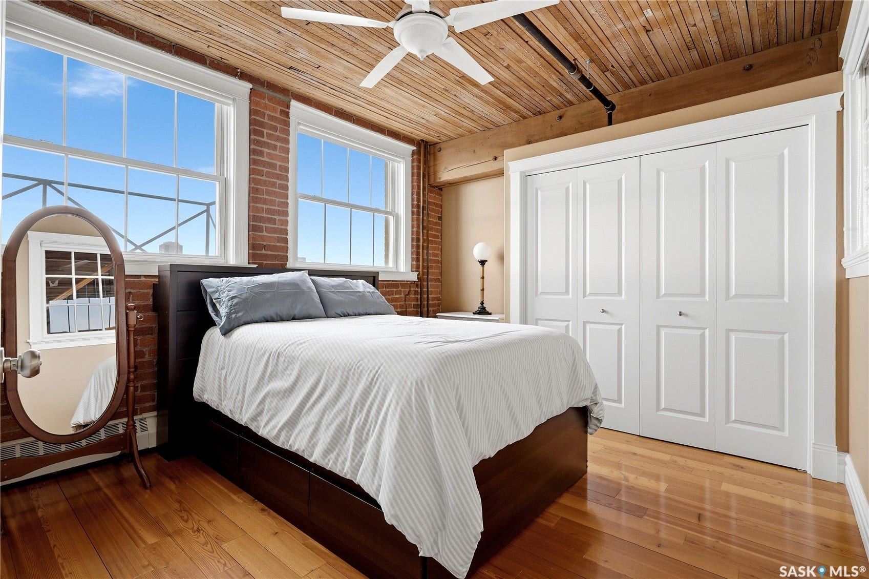 Exposed brick wall, two large windows, wood-finish flooring, wood plank ceiling, and white bifold closet doors