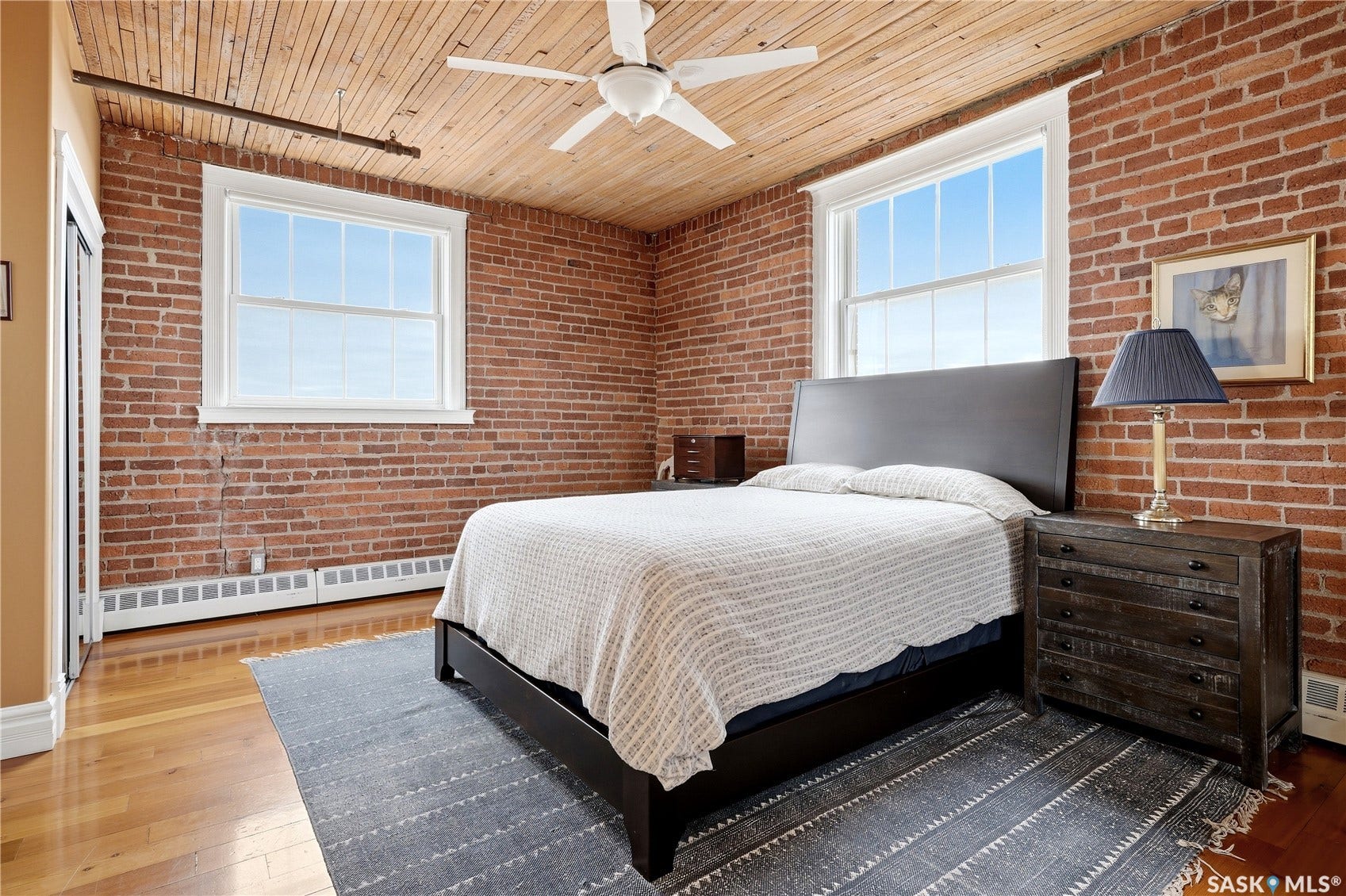 Exposed brick walls, wood-paneled ceiling, wood-finish flooring, two large windows, and a ceiling fan