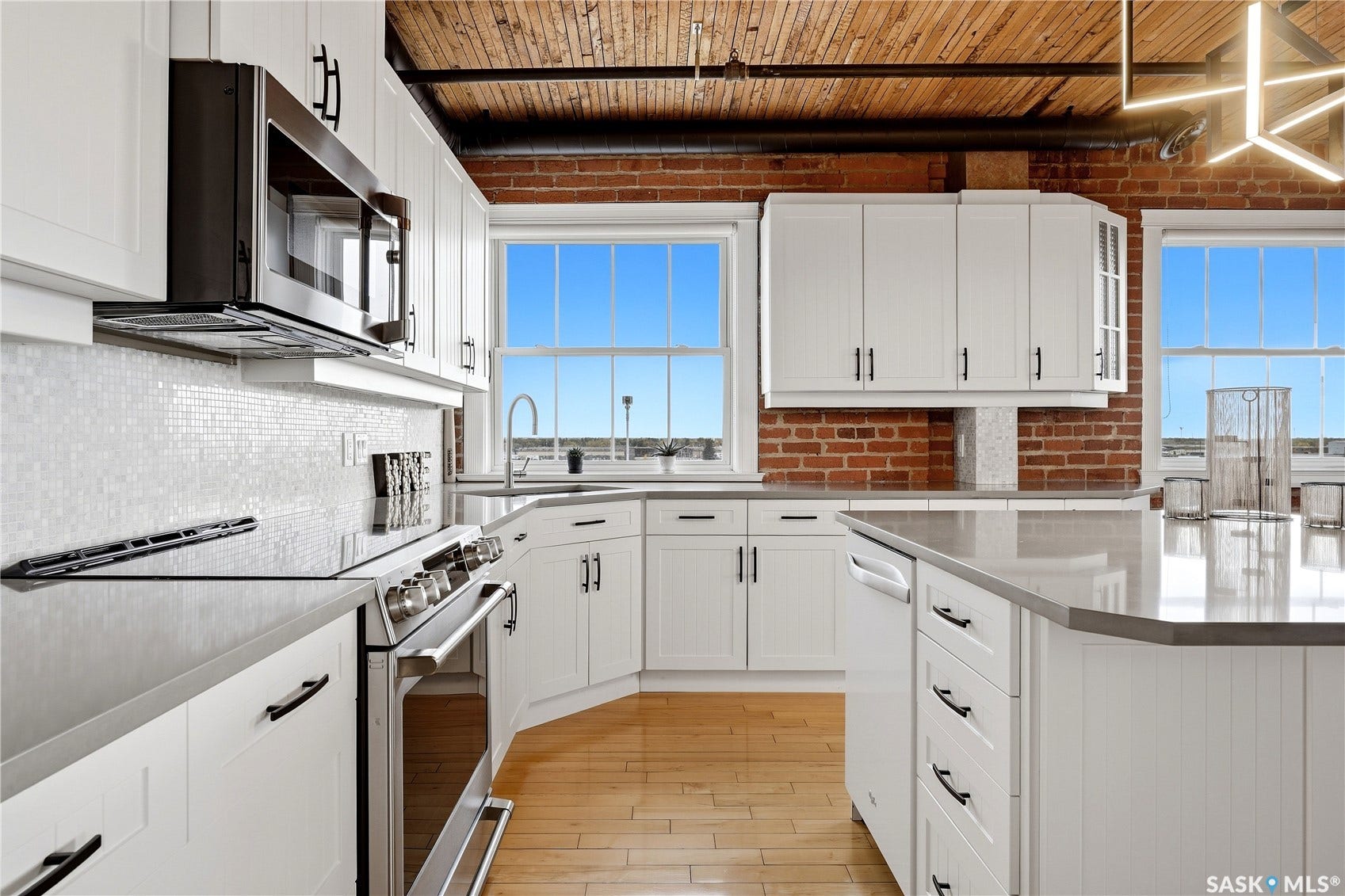 White cabinetry, exposed brick accents, stainless steel appliances, wood-finish flooring, and a large central island with solid surface countertops