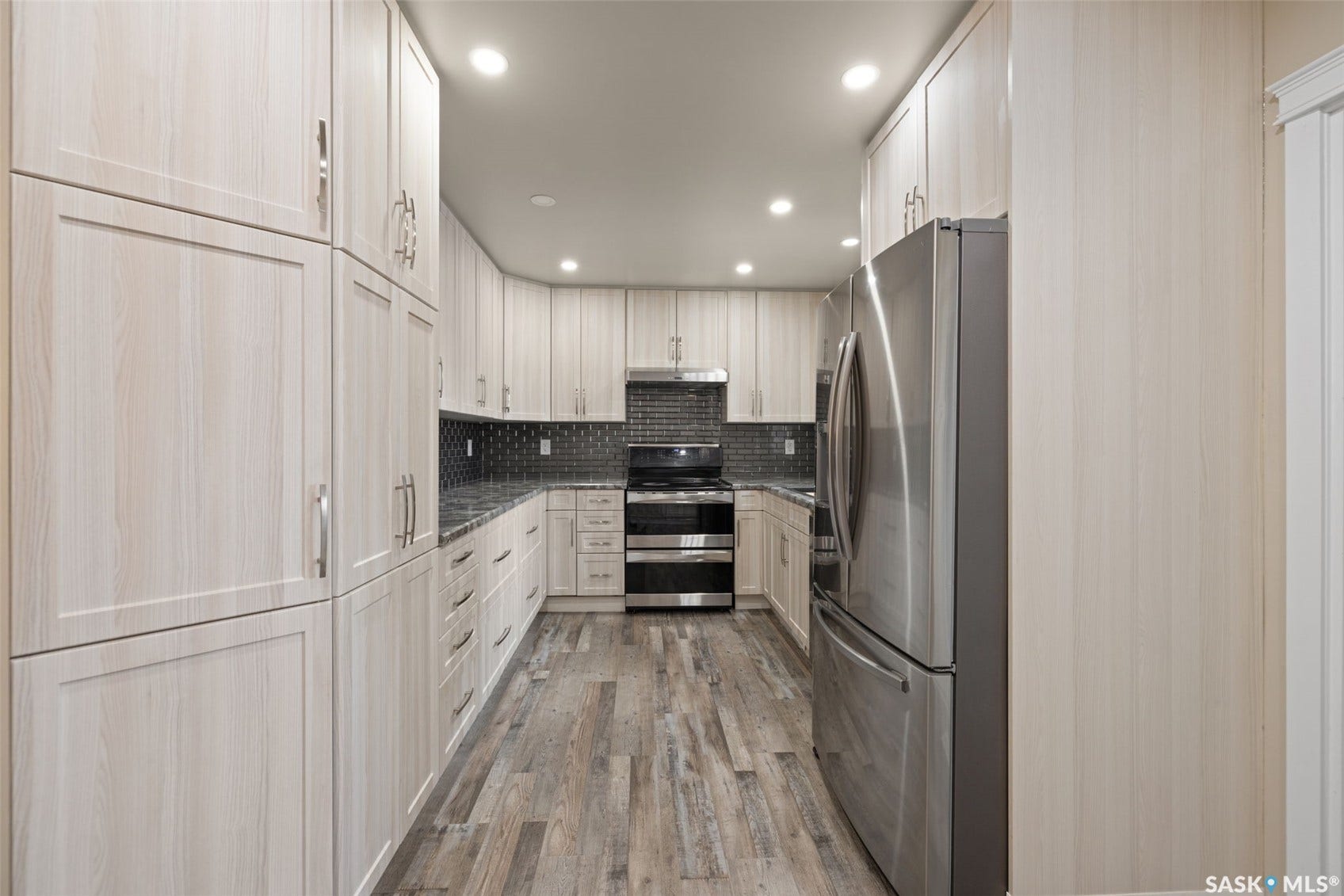 Kitchen featuring extensive light wood-finish cabinetry, stainless steel appliances, dark tile backsplash, dark countertops, and wood-finish flooring